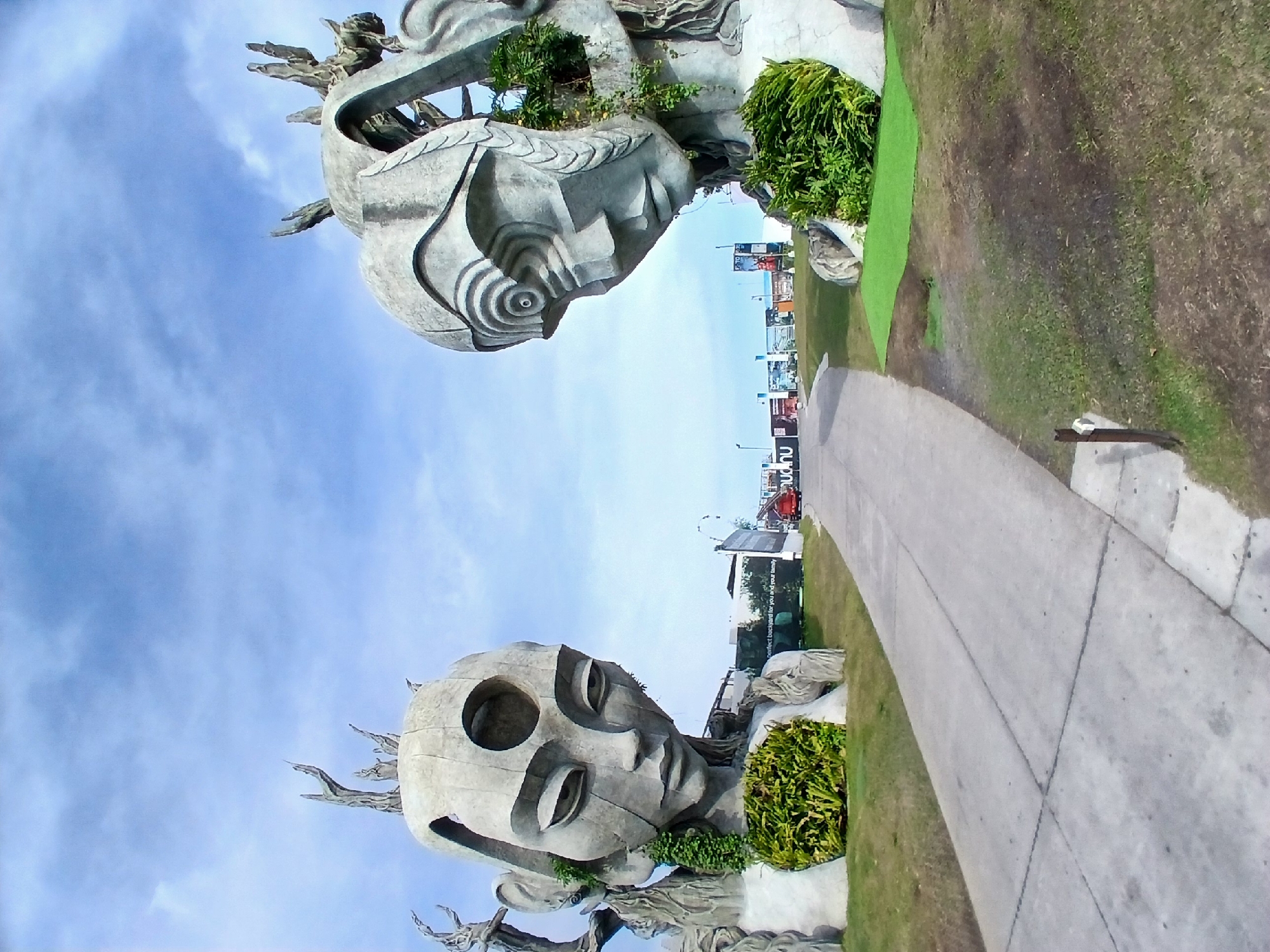 This is a wide-angle, eye-level outdoor photograph of two large, abstract sculptures resembling human heads, located in front of buildings under a partly cloudy blue sky. The sculptures are made of a grey, stone-like material and are positioned on either side of a paved walkway leading into the distance. The sculpture on the left is larger and more detailed, with a distinctly feminine face, a circular opening in the forehead, and vegetation growing from its chest cavity. The sculpture on the right is partially obscured and appears more stylized, with a complex, layered design around the eye area and vegetation growing within its structure. The walkway is made of concrete slabs and is flanked by green grass and some large rocks. In the background, there are several signs and banners, and a red truck is visible on the walkway. This scene is part of the Kediri Old Town cultural complex in Kediri, Indonesia, a popular tourist attraction known for its artistic installations and historical significance.