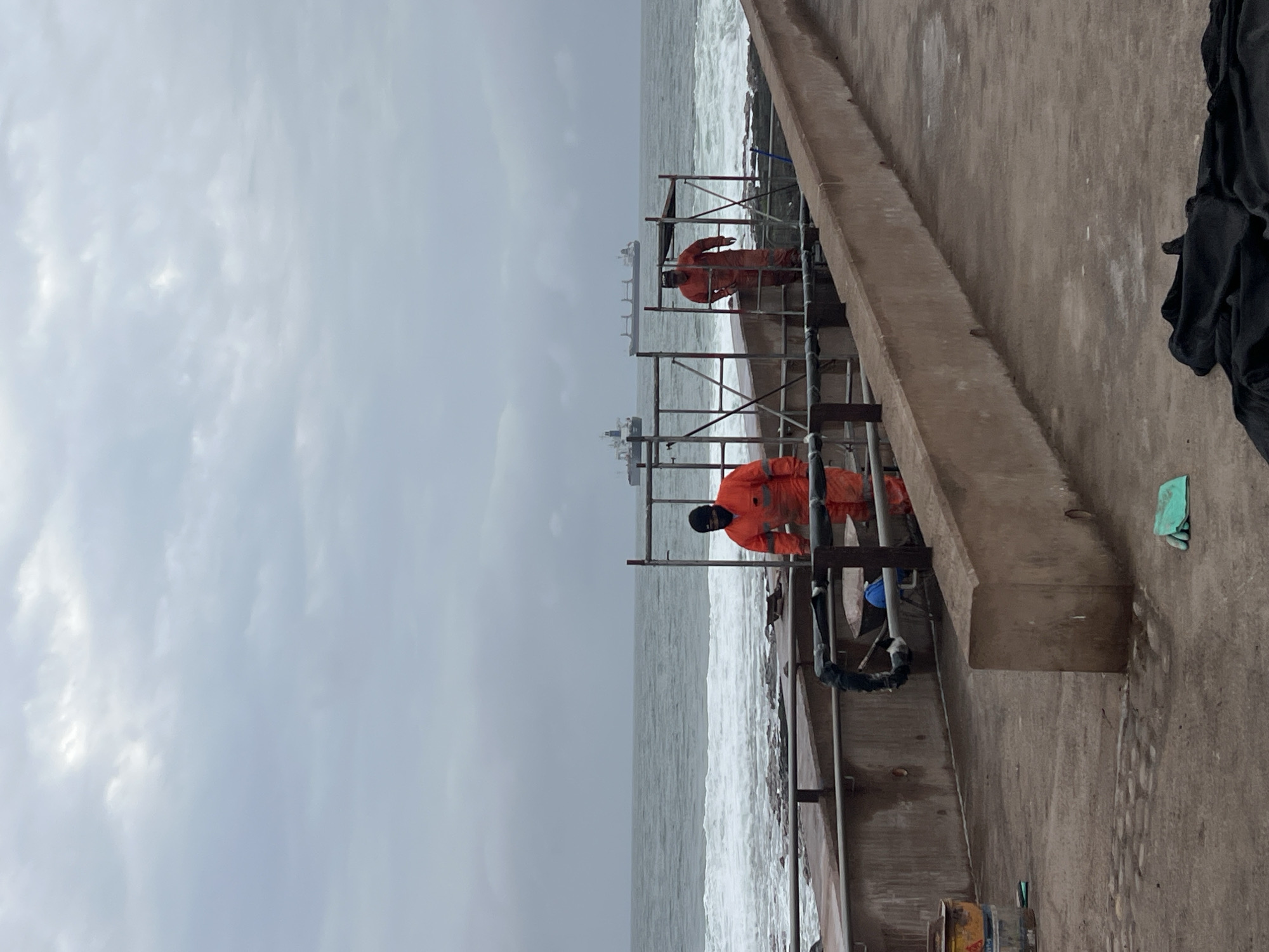 The image depicts a scene by the ocean, possibly a coastal promenade or construction site in Arica, Chile, under an overcast sky. In the foreground, a concrete structure runs diagonally across the frame. Two individuals are visible on a raised platform, both wearing bright orange protective suits and face coverings. One individual, closer to the viewer, appears to be walking towards the right, while the other is positioned further back, nearer to the water. Scaffolding is erected around the platform where the individuals are working.
Beyond the platform, the ocean stretches out, with moderate waves breaking on a rocky shoreline. Two cargo ships are visible in the distance on the horizon. In the immediate foreground, on the concrete surface, there are various objects including yellow and blue buckets, a blue object that might be a helmet, some black fabric, and a pair of green gloves. The overall atmosphere is subdued, likely due to the cloudy weather. The presence of scaffolding, protective gear, and what appear to be construction materials suggests ongoing work at this coastal location.