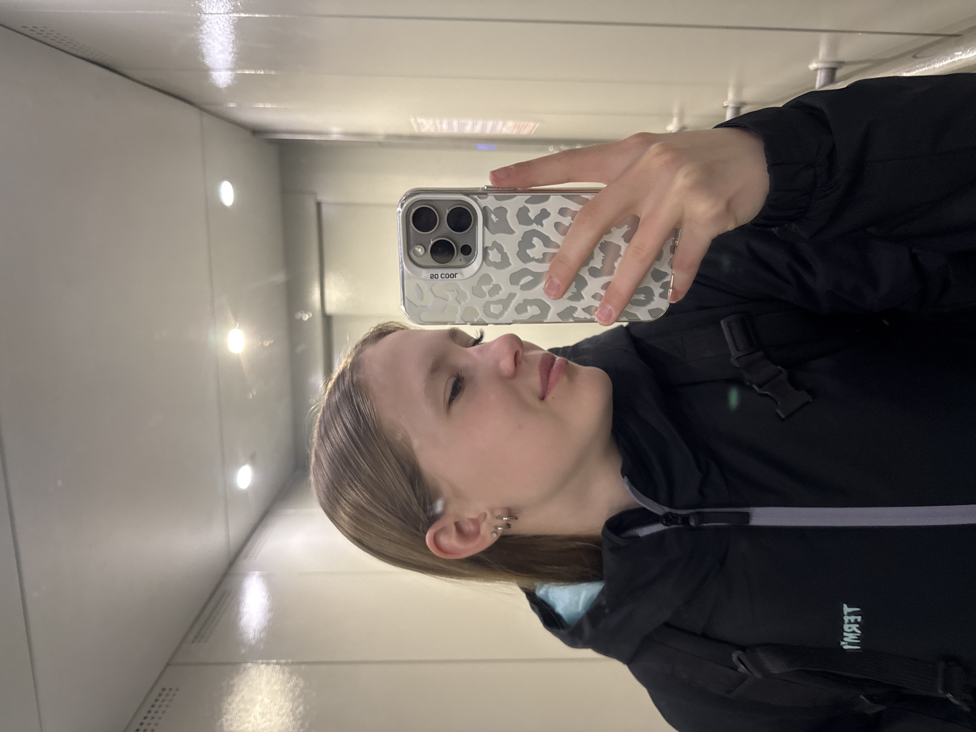 A young woman is captured in a mirror selfie within an indoor setting, likely a corridor or elevator, in Chaikovsky, Russia. The environment features uniform, light-colored, reflective walls and ceiling, illuminated by several bright, embedded circular lights.
The woman is shown in profile, looking slightly upwards with her eyes partially closed, conveying a serene or contemplative expression. She has light brown hair and is wearing a black jacket, possibly a puffer or windbreaker, with a light blue lining visible near the collar. A black backpack strap is visible over her right shoulder. Her left ear features multiple metallic hoop piercings.
She holds a smartphone in her right hand, which is fitted with a distinctive grey and white leopard-print case. Text visible on the phone case, as seen in the reflection, reads "2O COOR". Additional text, "ТЕРМТ", is visible in reflection on her jacket, suggesting a brand name. The image captures a casual moment in a well-lit, modern-looking indoor space.
