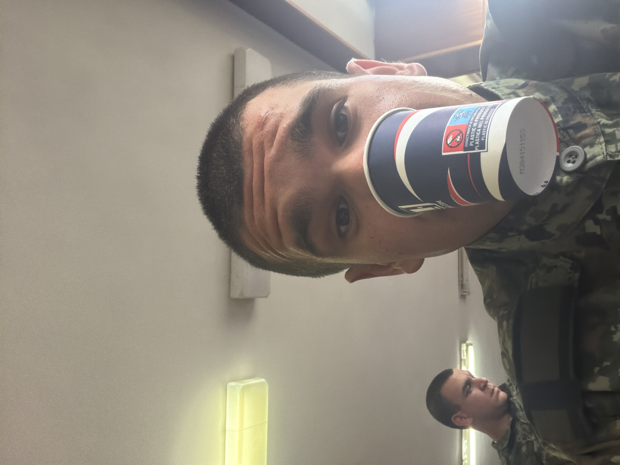 The image, rotated 90 degrees clockwise, captures two individuals indoors. In the foreground, a man with short dark hair and dark eyes is shown from the chest up, his head tilted towards the viewer. He appears to be wearing a camouflage uniform, with a visible shirt button. He holds a dark blue paper cup, featuring red and white stripes, directly to his face, partially obscuring his mouth and nose. His expression is neutral, showing slight forehead wrinkles.
The paper cup displays several pieces of text and symbols. Visible text includes "ПЛАСТМАСА В ПРОДУКТ" (Bulgarian for "PLASTIC IN PRODUCT"), "PLASTIC IN PRODUCT" (English), "PLASTICA NEL PRODOTTO" (Italian for "PLASTIC IN PRODUCT"), and "PLÁSTICA EN PRODUTO" (likely Portuguese or Spanish for "PLASTIC IN PRODUCT"). A recycling symbol with a crossed-out person, indicating "do not litter," is also visible. A numerical code, "M304151153," is printed on the cup's bottom rim, and the partial word "COM" is on its side.
In the background, a second man, also in a camouflage uniform, is partially visible in profile. He has short dark hair and appears to be looking to his left with a serious or pensive expression.
The setting is an indoor space with plain, light-colored walls. Artificial lighting, including fluorescent tubes and a rectangular fixture, illuminates the scene. The uniforms suggest a military or similar service environment, possibly a barracks or training facility in Ledenik, Bulgaria. The activity seems to be a casual moment, with the main subject possibly drinking from or holding the cup. The time of day is indeterminate due to the artificial lighting.