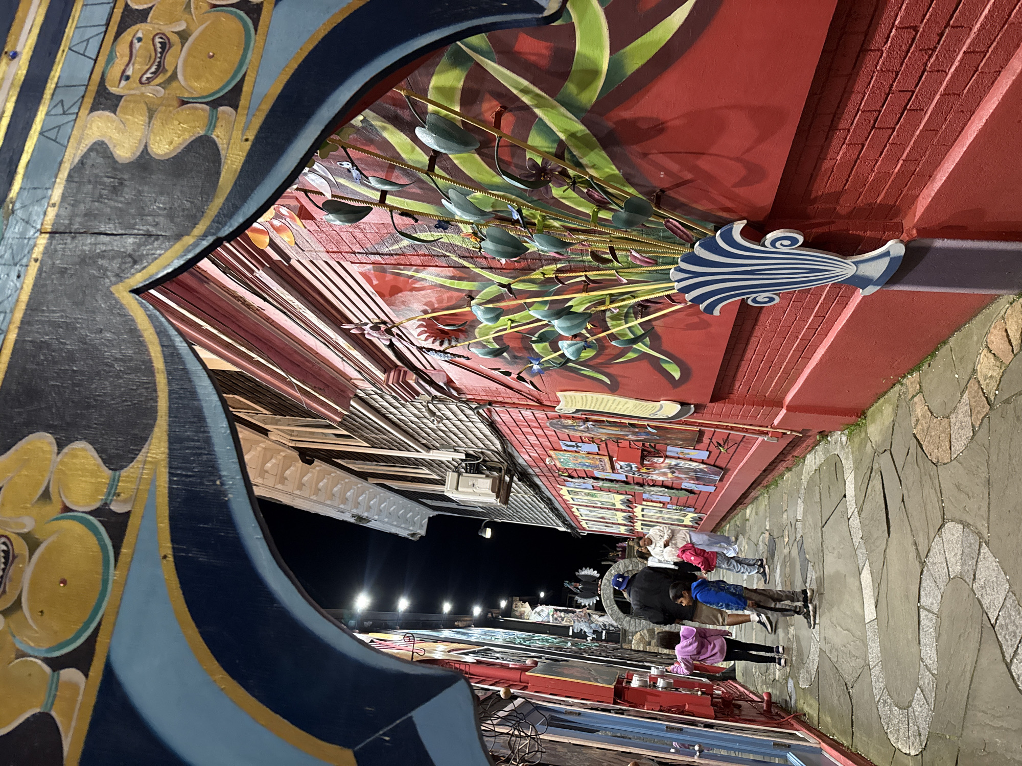 The image shows a pedestrian street in Provincetown, USA, viewed through an elaborately decorated archway. The archway is painted with stylized floral and geometric patterns in black, gold, blue, and other colors. It frames a street lined with buildings, many with brightly colored facades and displaying artwork. In the middle of the street, several people are walking away from the viewer, including individuals wearing a pink jacket, a blue shirt, and a white top. The street is paved with cobblestones or patterned stones. The buildings are of varying heights and styles, and some have decorative elements. Above the buildings, streetlights provide illumination. The overall atmosphere is colorful and lively.