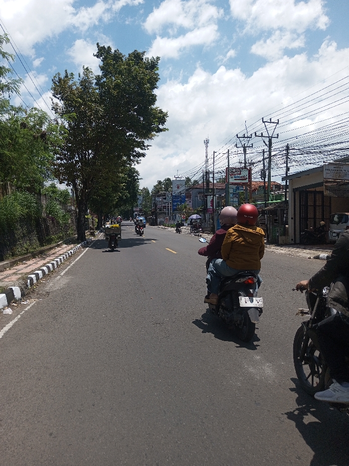 The image depicts a daytime street scene in the city of Tagtse Tashidingkha, Bhutan, under a blue sky with scattered white cumulus clouds, casting distinct shadows on the asphalt road. The road, appearing worn, features faint yellow dashed and solid line markings. On the left, a concrete curb with alternating black and white stripes borders a vegetated embankment populated by mature trees and dense foliage.
On the right side of the road, a series of multi-story buildings with various commercial signage, including a partially visible "Alfamart" logo, line the street. Numerous utility poles supporting a dense network of power lines stand in front of these structures, and a tall cellular tower is visible in the distance.
In the mid-ground, a dark scooter with two occupants is moving away from the viewer. The driver wears a maroon jacket and a light pink helmet, while the passenger wears a mustard yellow jacket and a red helmet, both individuals wearing blue jeans. The scooter's rear license plate is clearly visible, displaying "F 3105 WAS 02.26." In the immediate foreground on the right, the front wheel, a partial dark frame, and the right arm and leg of a rider on another dark motorcycle are visible, also moving forward. Further down the road, additional motorcycles and scooters are present, one on the left carrying a large package. Several pedestrians are visible near the commercial buildings on the right.
