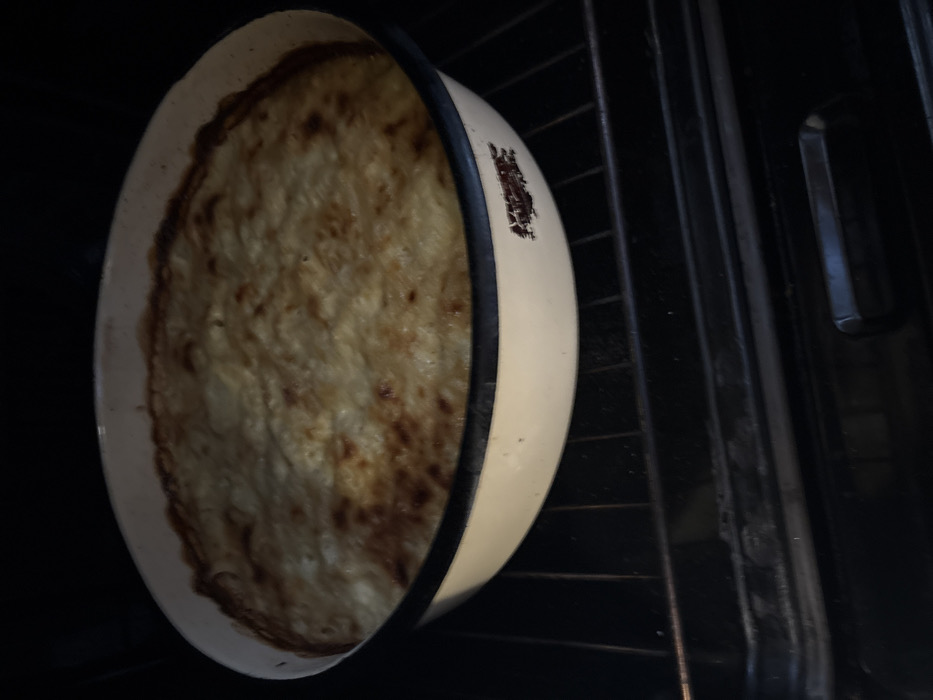 A round, cream-colored enamel baking dish, approximately 30-35 centimeters in diameter, occupies the center of the frame, resting on a black metal oven rack. The dish contains a baked food item with an unevenly browned, golden-tan surface, indicative of a gratin or casserole. The texture of the food appears soft and possibly creamy, with darker, crisped edges. The enamel dish features a dark rim and shows signs of wear, specifically a chipped area on its right side, revealing a reddish-brown subsurface material. The interior of the oven, characterized by its dark, metallic surfaces and additional lower racks, serves as the environment. No individuals, overt actions, or interactions are present in the image. The depicted scene is located in the city of Stefan Karadja, Bulgaria.