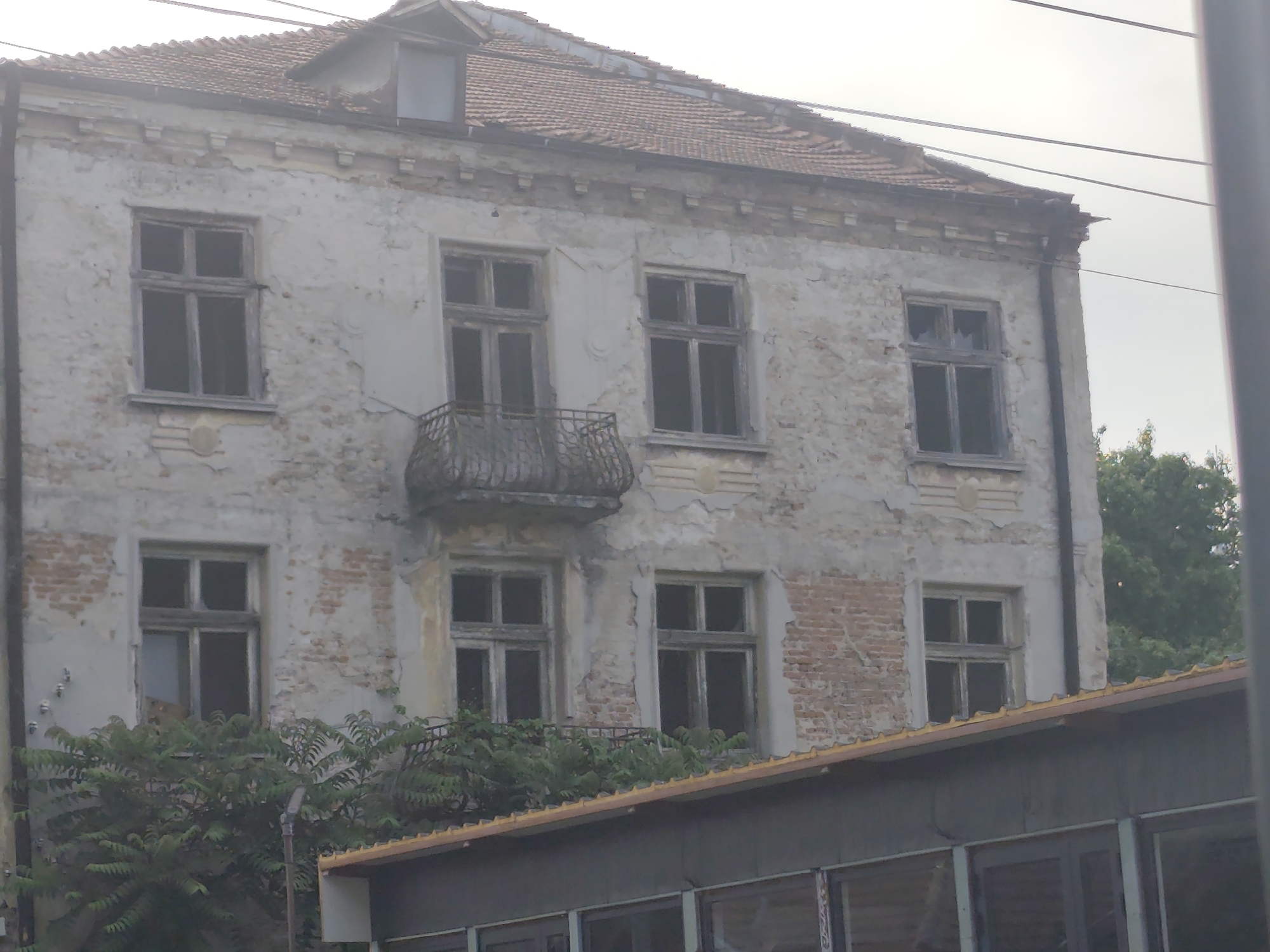 This image depicts a two-story residential building in a state of advanced disrepair, located in Lom, Bulgaria. The facade is characterized by severely deteriorated light-colored plaster, which has extensively peeled and crumbled, exposing sections of red brick masonry beneath. The building features multiple rectangular window openings on both visible floors, fitted with multi-pane wooden frames. The window panes appear dark or absent, suggesting either broken glass or shadowed interiors. Centrally located on the second floor is an ornate, curved wrought-iron balcony. The roof is composed of red ceramic tiles and includes a gabled dormer window with a single pane near the peak. A deteriorated cornice and decorative band are visible below the roofline. Dense green foliage partially obscures the lower facade of the main building and the roofline of a lower, separate structure in the immediate foreground. This foreground structure has a low-pitched roof with a light-colored fascia and appears to have large windows, indicating a different architectural style or function. Overhead utility lines are visible traversing the upper portion of the frame against a pale, overcast sky. A dark vertical column, possibly a utility pole or building edge, frames the right side of the image. No individuals or discernible activity are present within the scene.