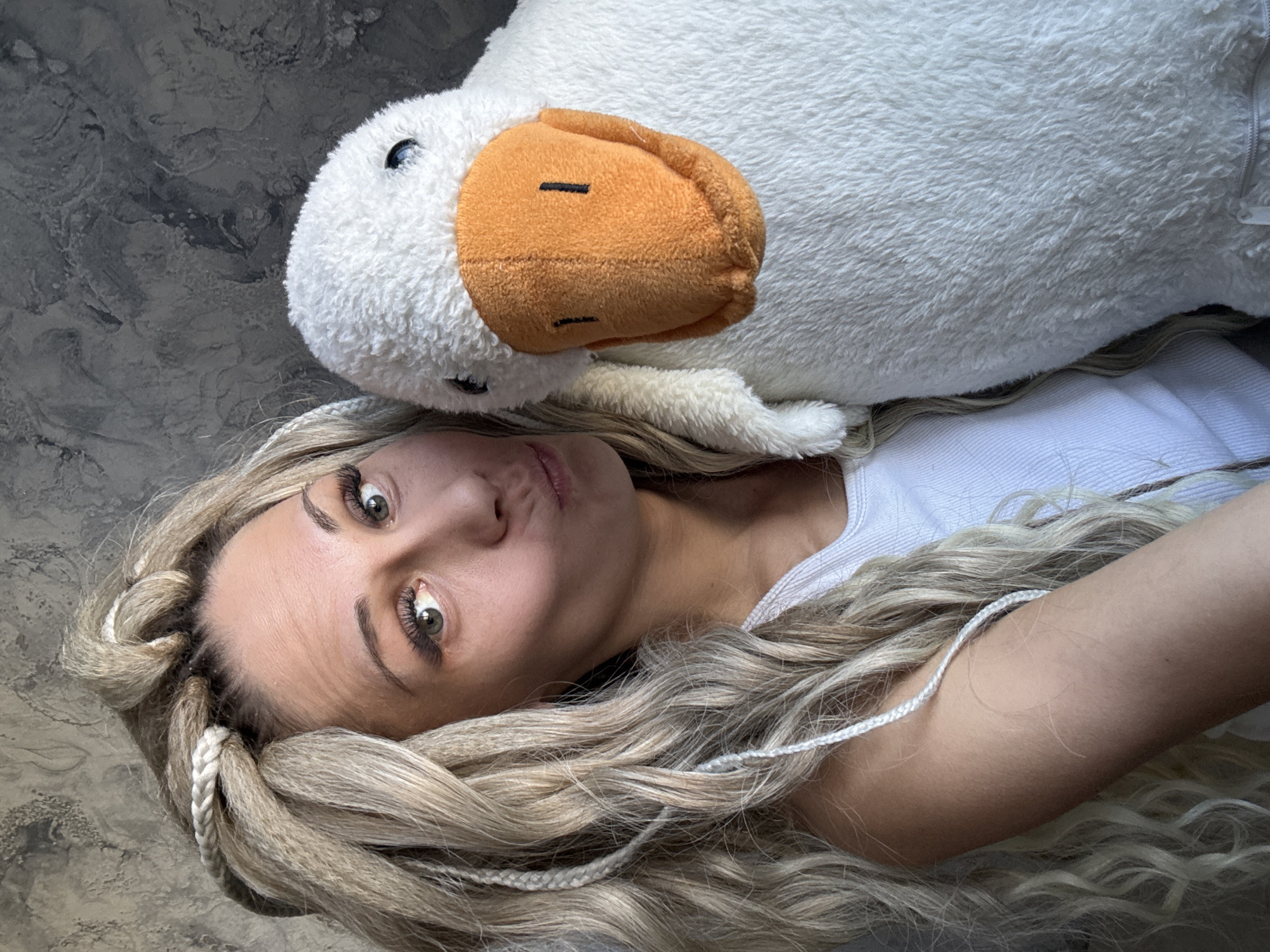 A woman is lying on a gray surface. She has blonde hair styled in braids and is wearing a white tank top. A large, stuffed white duck with an orange beak is lying on top of her. The background appears to be a mottled gray, possibly a wall or floor. The location is unknown. There is no information to indicate that the location is in Rusanovo, Russia.