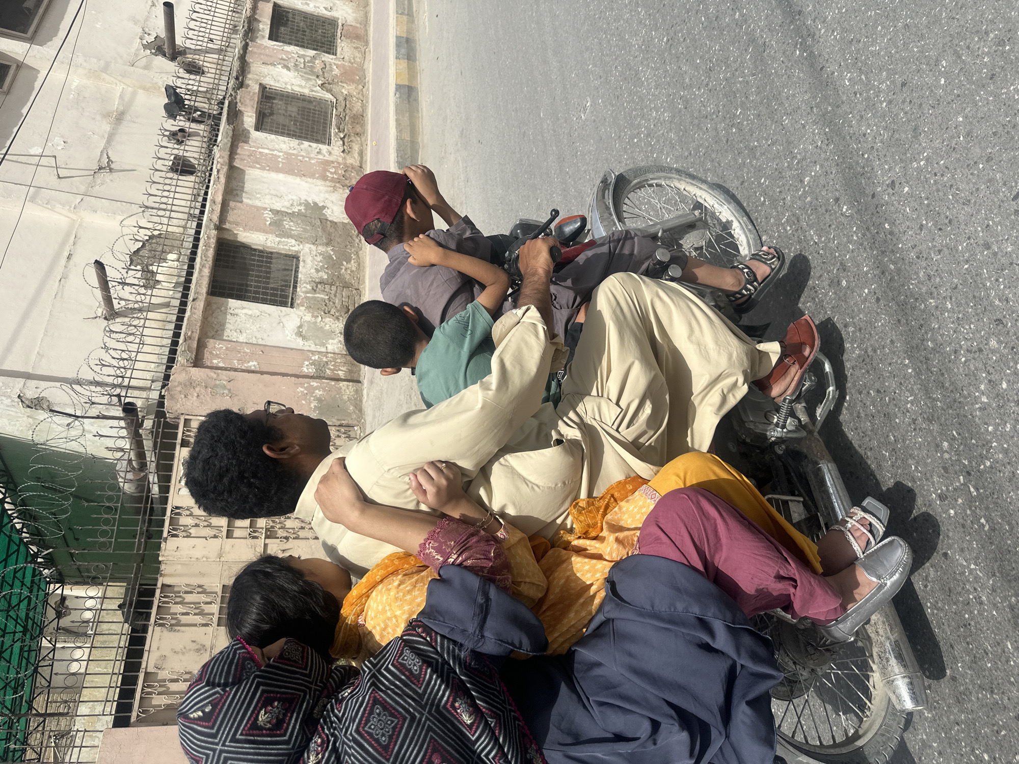 In Quetta City Tehsil, Pakistan, a family of five is riding on a motorcycle. A man is driving, with a woman and two children seated behind him, and a third child is perched on the back. The family is dressed in traditional clothing. The motorcycle is on a paved road, with buildings and a barbed-wire fence visible in the background. The sun is shining, casting shadows on the ground.
