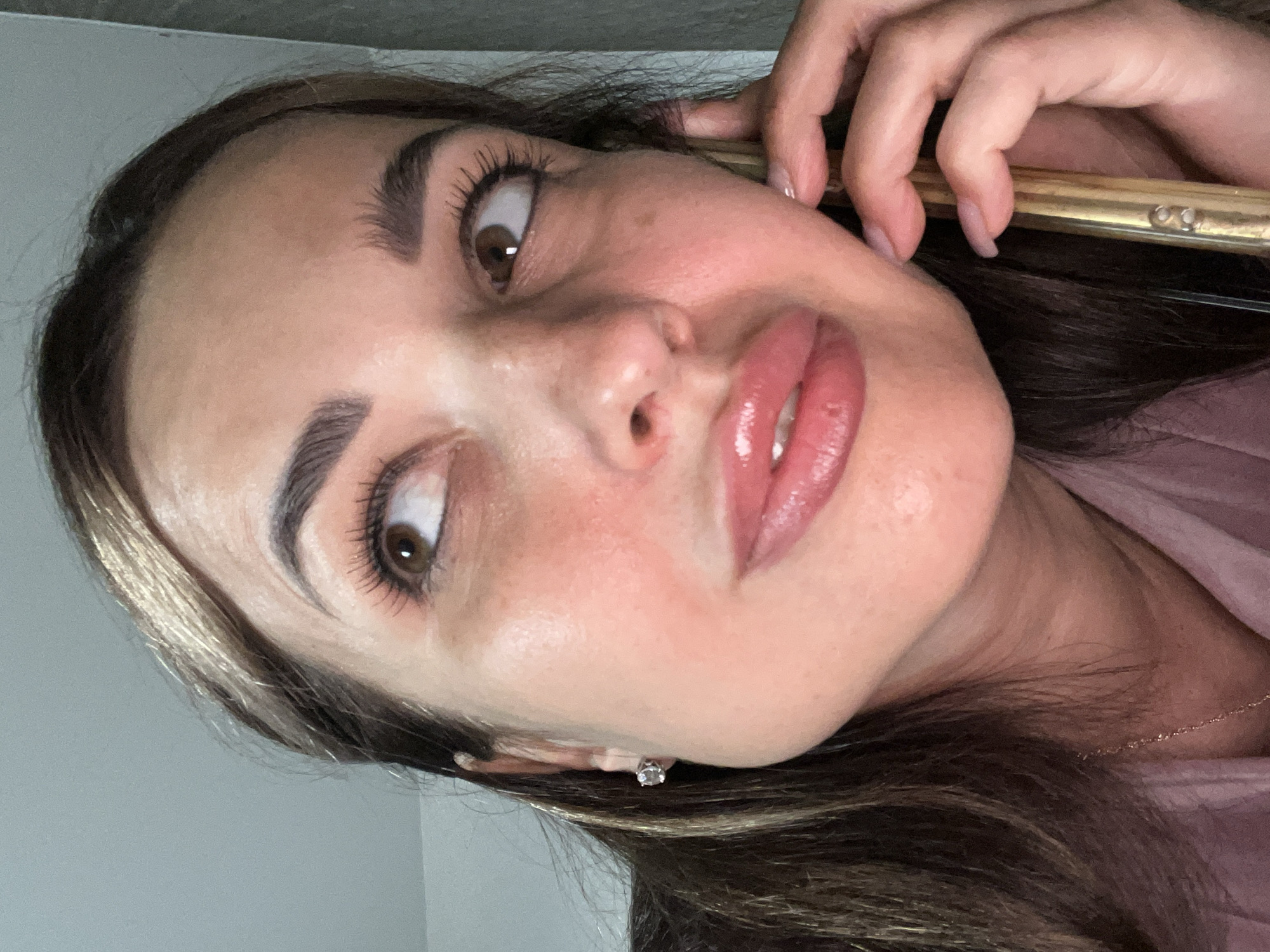 A woman is seen from the shoulders up, in a close-up shot. She has brown hair, brown eyes, and light skin. She is wearing a pink shirt and a gold necklace. She is holding a gold cell phone to her right ear and appears to be looking up and to her left. Her lips are parted, and she has on a glossy lip product. There is a diamond stud earring in her left ear. The background is a neutral gray.