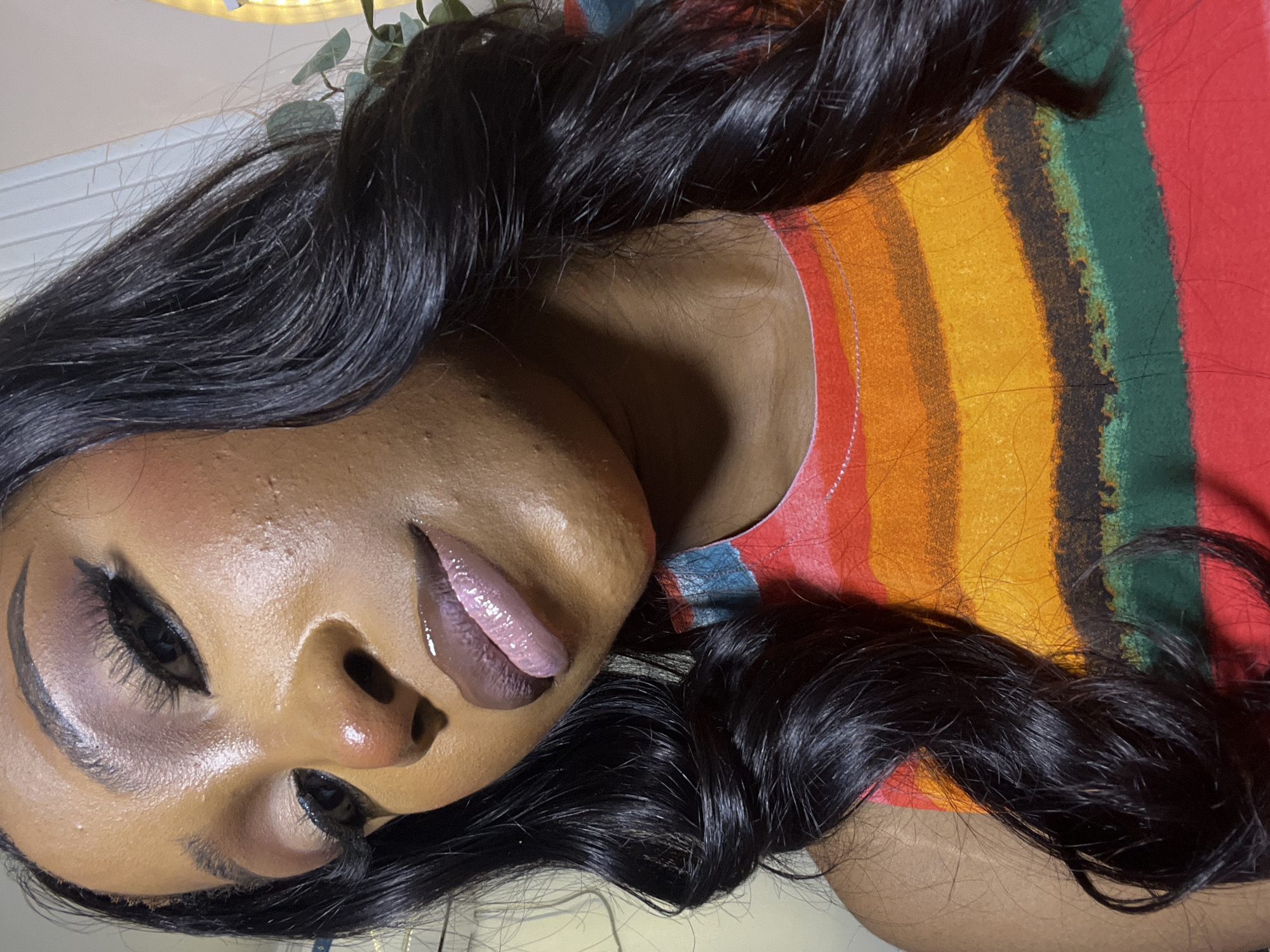 The image shows a close-up, waist-up shot of a young Black woman with dark, wavy hair. She is wearing a colorful, striped t-shirt with red, orange, yellow, green, and black bands. The woman is looking down and slightly to the left, with her makeup on. She has on eyeshadow, eyeliner, mascara, blush, and lip gloss. Her skin has a dewy finish, and there are some small blemishes visible on her cheeks and nose. The background is out of focus and appears to be an indoor setting, with a glimpse of a plant on the right. The lighting is warm and highlights the subject's features. There is no discernible indication of the city of Abuja, Nigeria, or any specific location within it.