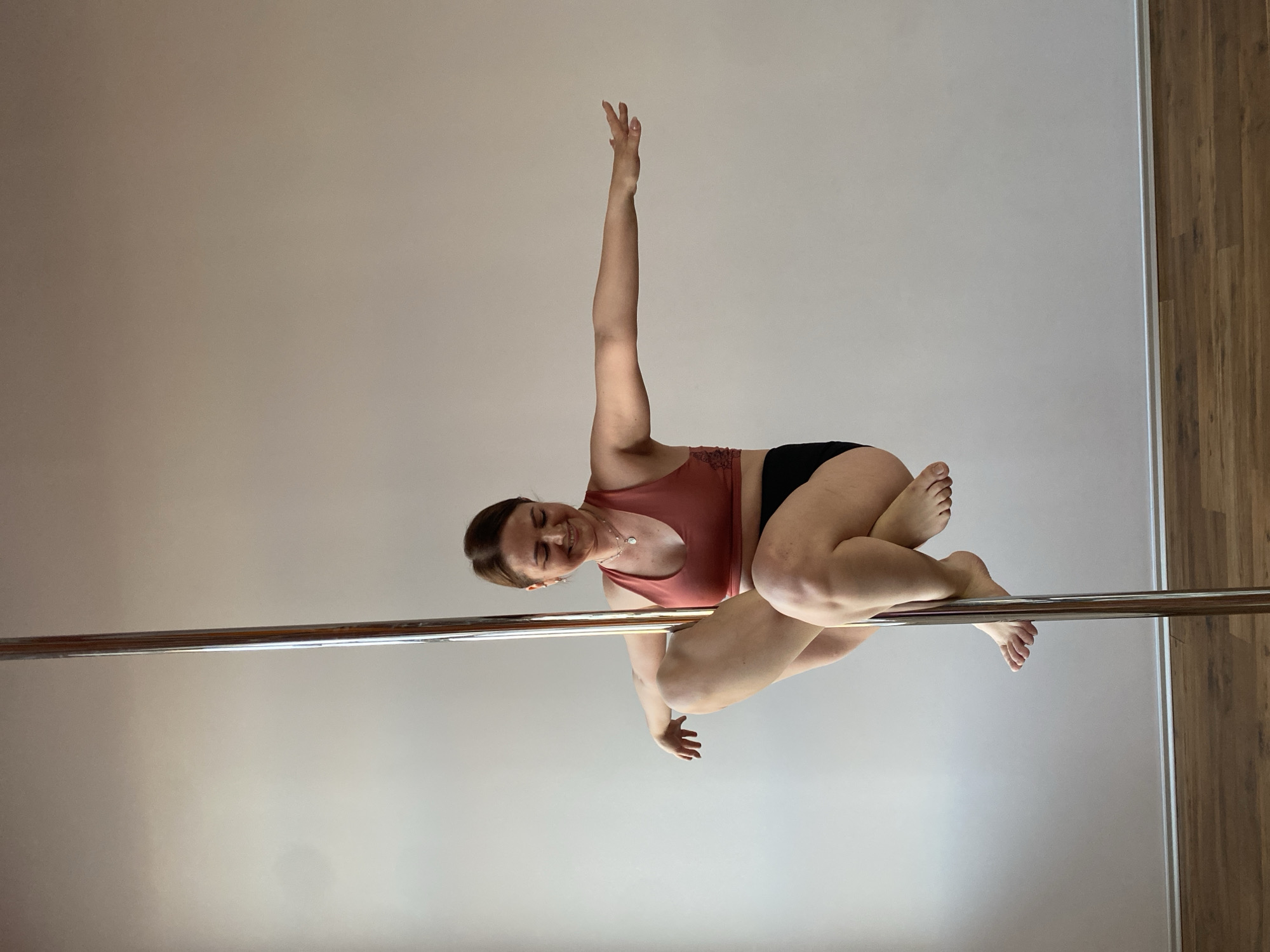 A woman is performing pole dancing in an indoor setting. She is gripping a horizontal metal pole with her legs and one arm while extending her other arm upwards. She is wearing a coral-colored sports bra and black shorts. The background consists of a light-colored wall and a wooden floor. The location is presumed to be Yekaterinburg, Russia, based on the prompt instructions, although no specific details in the image confirm this.