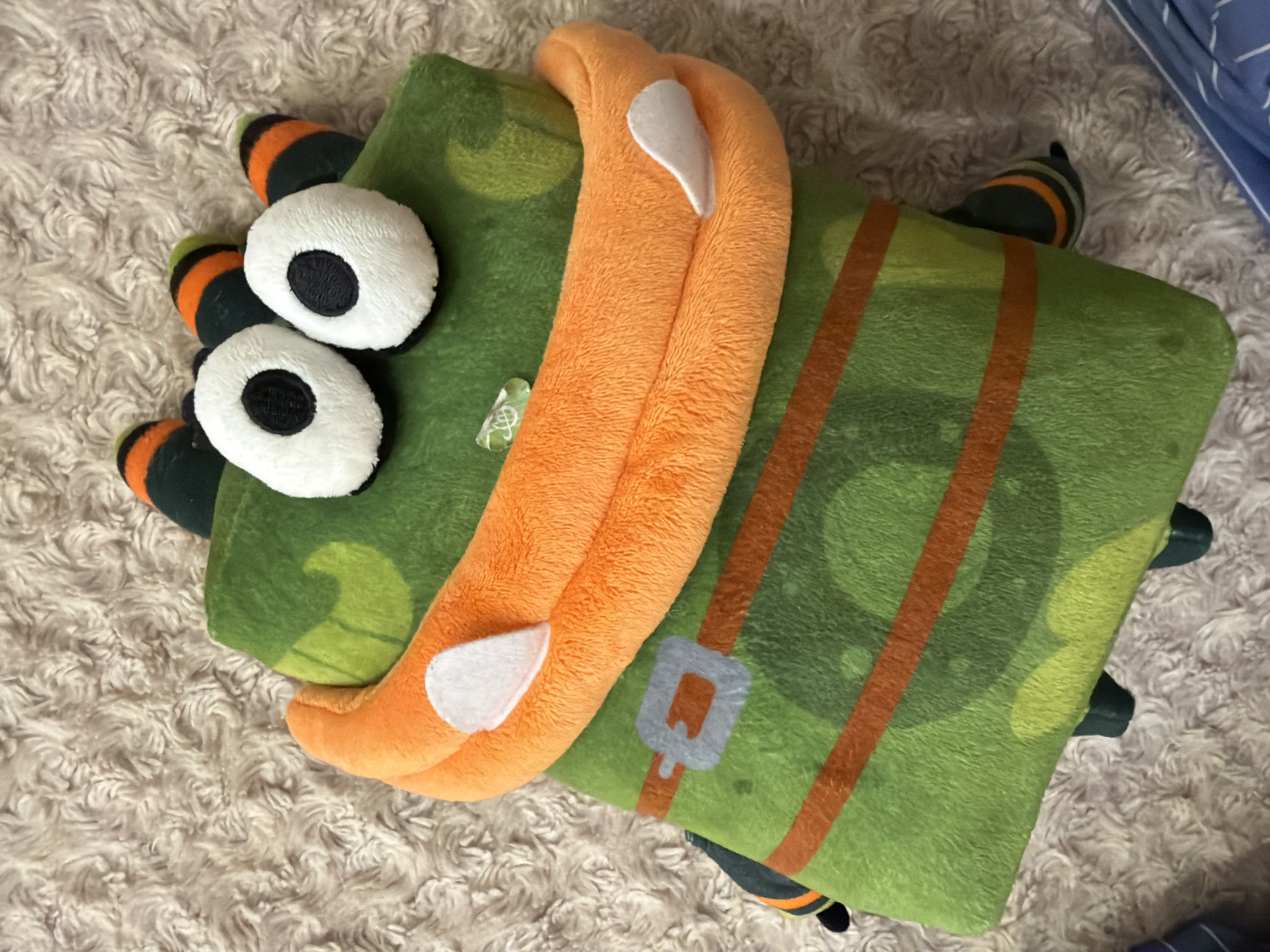 This media file shows a plush toy lying on a fluffy, cream-colored rug. The plush toy is green and rectangular, with orange stripes that resemble suspenders and a light blue buckle. It has two large, white, oval eyes with black centers, giving it a cartoonish appearance. The toy also has several black, orange, and green striped appendages sticking out from its sides and head, which could be interpreted as legs or fins. An orange plush element, shaped like a mouth or a smile, is attached to the front of the toy, with two white felt-like shapes as accents, possibly representing teeth or highlights. The lighting suggests it is daytime, and the indoor setting is indicated by the rug and the presence of a blue patterned fabric, possibly bedding, in the background. There are no visible text elements in the image. The overall impression is that of a playful, brightly colored toy.