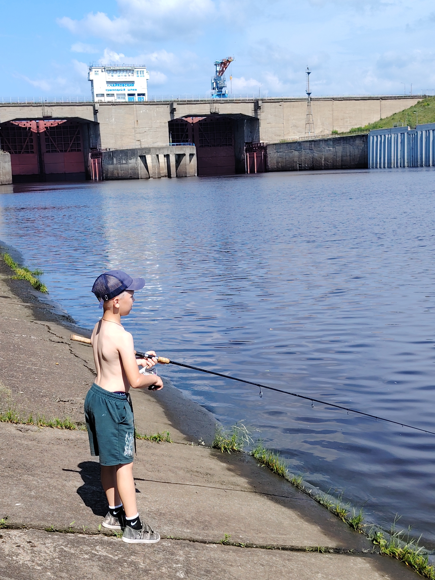 A boy is fishing from a concrete embankment on the Kama River in Chaikovsky, Russia. The boy is wearing a blue baseball cap, dark green shorts, and camouflage sneakers with white socks. He is holding a fishing rod, casting into the blue water. In the background, a large dam structure is visible, with a building on top labeled "г. ЧАЙКОВСКИЙ судоходный шлюз" (Chaikovsky, Shipping Lock). The sky is blue with scattered white clouds. The sun is shining, casting shadows.