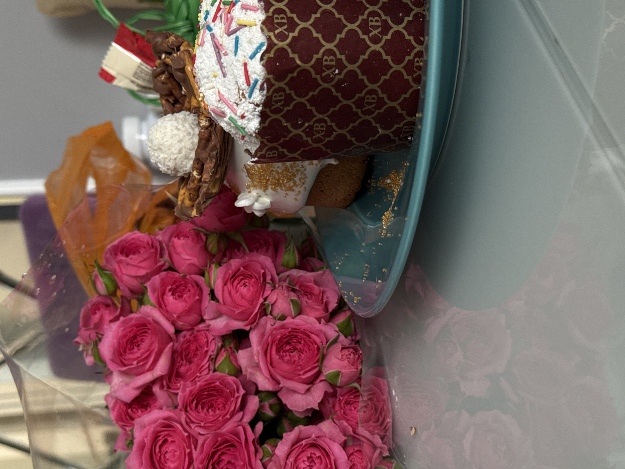 The image shows a close-up of an Easter cake decorated with white frosting, colorful sprinkles, and chocolate adornments, presented alongside a bouquet of pink roses. The cake is partially encased in a decorative maroon and gold patterned paper. It sits on a light blue dish or plate, speckled with gold dust. The roses are wrapped in clear plastic. The setting appears to be indoors, with a reflective surface behind the flowers and cake, possibly a wall or glass. The location is not specified and is labeled as Unknown, Unknown.
