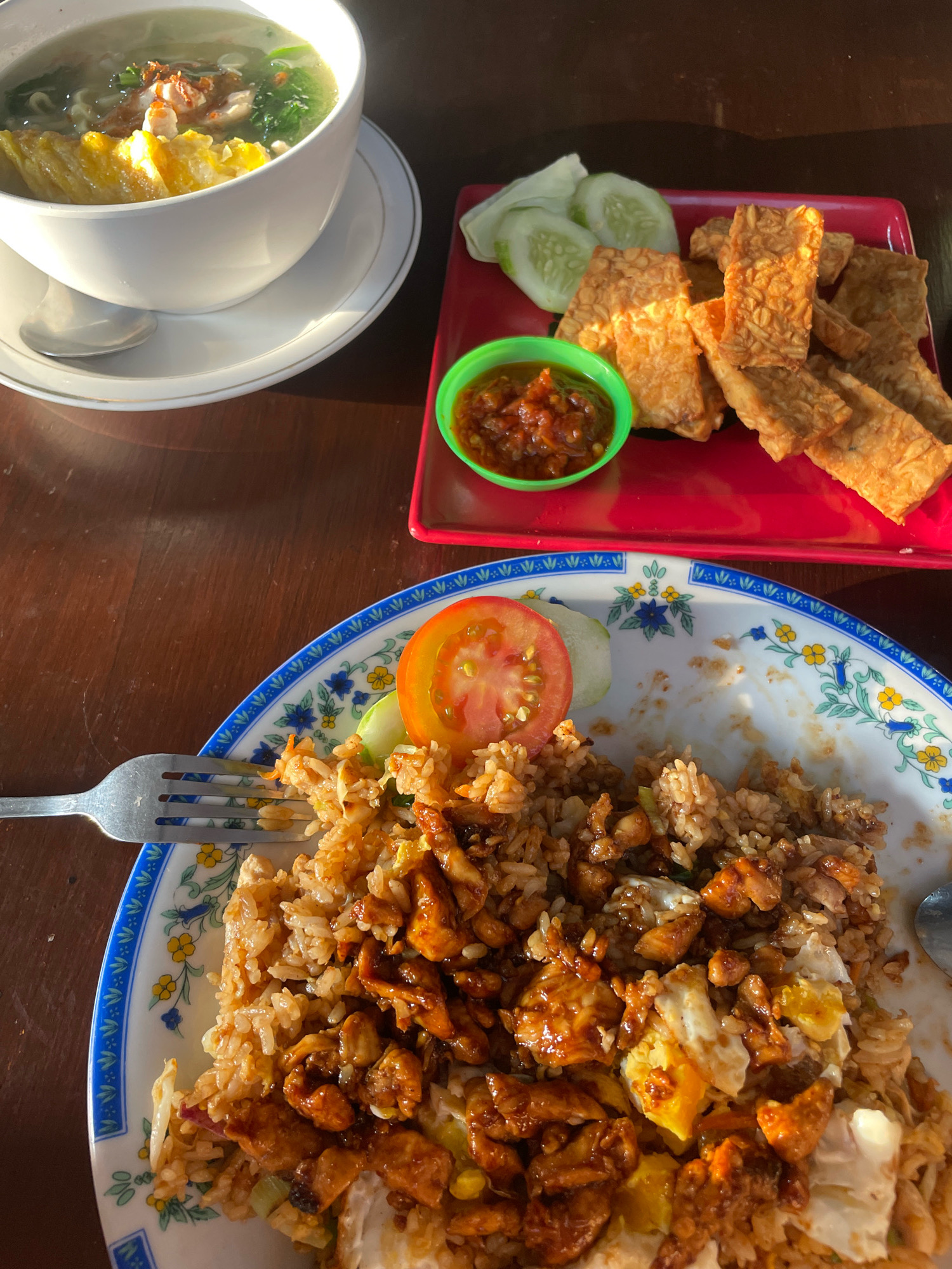 An overhead shot captures a meal served on a wooden table in Kuta Selatan, Indonesia. In the foreground, a white plate with a blue and white floral rim holds a generous portion of fried rice, interspersed with chunks of chicken and scrambled egg. A fork rests to the left of the plate, and a spoon is visible on the right. Behind the fried rice, a red rectangular plate contains several pieces of fried tempeh, sliced cucumber, and a small green bowl filled with a reddish condiment, likely sambal. To the left, a white bowl on a saucer contains soup with noodles, egg, and some greens. The lighting suggests it is daytime, possibly late afternoon given the warm tones and shadows.