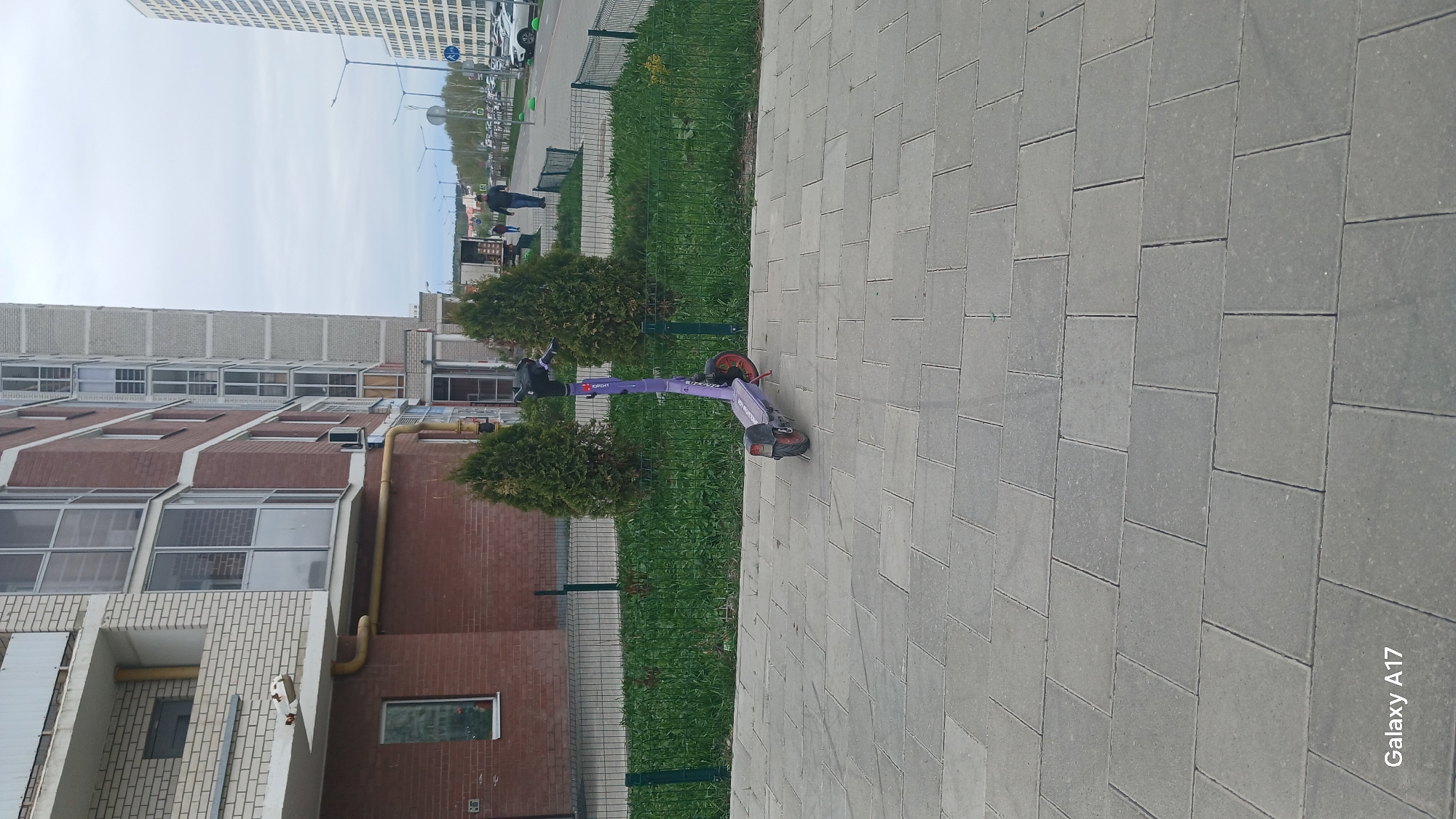 The scene shows a purple electric scooter parked on a gray tiled sidewalk in Yekaterinburg, Russia. The scooter is near a green metal fence and two small evergreen bushes. Behind the fence is a grassy area next to a multi-story residential building constructed of brick and concrete. In the distance, other buildings, streetlights, and pedestrian activity are visible. The sky is overcast. A watermark indicates the image was taken with a Galaxy A17 phone.