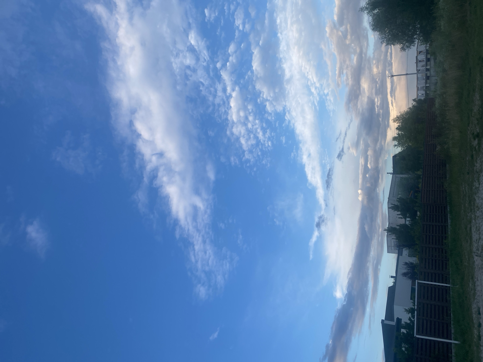 A bright blue sky with scattered white and gray clouds stretches above a residential area in Novi Petrivtsi, Ukraine. The foreground features a dark brown wooden fence with a white soccer goalpost on the left side. Beyond the fence, several modern houses with dark roofs are visible, interspersed with green trees. In the background, unfinished building structures and utility poles can be seen against the horizon. The overall scene depicts a suburban landscape under a partly cloudy sky.