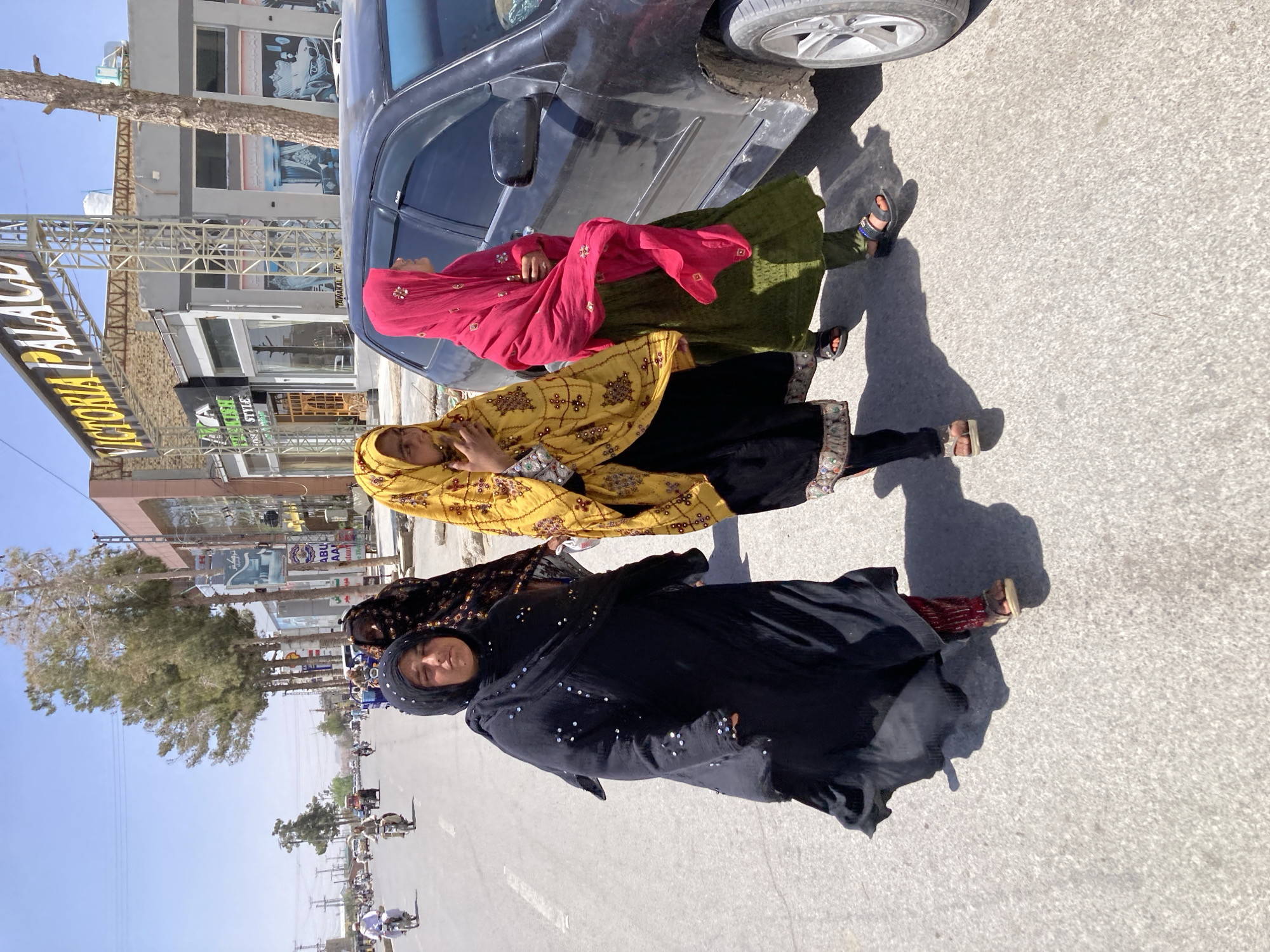 The image presents an outdoor scene on a sunny day in Quetta City Tehsil, Pakistan, featuring three women standing on the side of a bustling street. A black car is partially visible parked next to them on the right.
The women are dressed in traditional South Asian attire. The woman closest to the car wears a red headscarf adorned with golden embellishments and a green dress. Adjacent to her, another woman is clad in a bright yellow headscarf or shawl with detailed embroidery, paired with a dark outfit that includes ornate cuffs. The third woman, prominent in the foreground, wears a black hijab covering her head and a flowing black abaya or dress embellished with small, reflective silver sequins. She looks directly towards the camera with a neutral expression, while the other two women are looking slightly away or downwards. Their footwear consists of sandals or slippers.
The setting is an urban street lined with multi-story buildings, indicative of commercial establishments. A large yellow sign above one building clearly spells out "VICTORIA PALACE." Below it, another shop sign reads "FASHION STYLE." Other signs with local script are also visible, along with parts of scaffolding. Trees are planted along the roadside, and in the distance, more pedestrians and motorcycles can be seen on the road, suggesting a busy thoroughfare. The bright sunlight casts distinct shadows, indicating it is likely mid-morning or early afternoon on a clear day.
