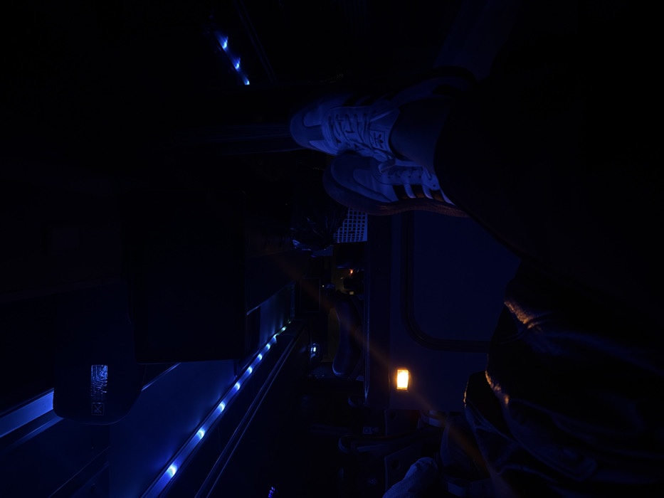 A person sits, legs crossed, in a dimly lit interior with blue lighting. The individual wears white Adidas sneakers. A metal safety barrier runs along the side. An orange light is visible in the background. The image suggests a confined space, possibly a train or vehicle interior at night.