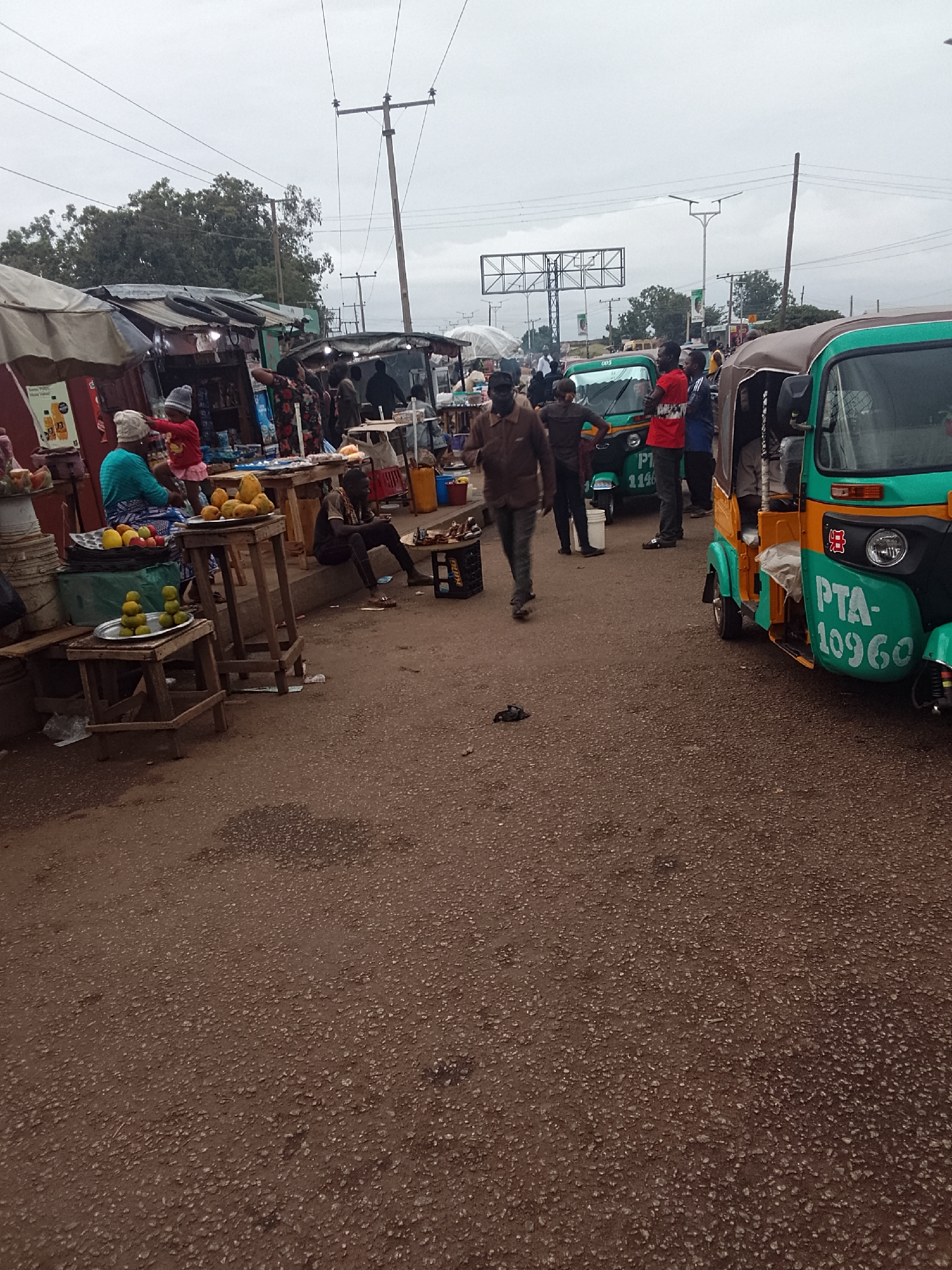 The image shows a street scene in Bukuru, Nigeria. The ground is brown and appears to be a road surface, possibly asphalt. On the left side of the image are stalls set up with vendors selling goods including fruit. People are present around these stalls, including one woman wearing a blue top and a child wearing a pink dress and white hat. A man sits on the side of the road using his phone. In the center of the image, a man in a brown jacket is walking towards the viewer. Further in the background, a group of men stand next to two green auto rickshaws, also known as "keke" or "tricycles", which serve as taxis. The license number of the auto rickshaw closest to the viewer is PTA-10960. Power lines and poles are visible above the street, indicating the presence of electricity. There is an overpass in the background. The sky is overcast.