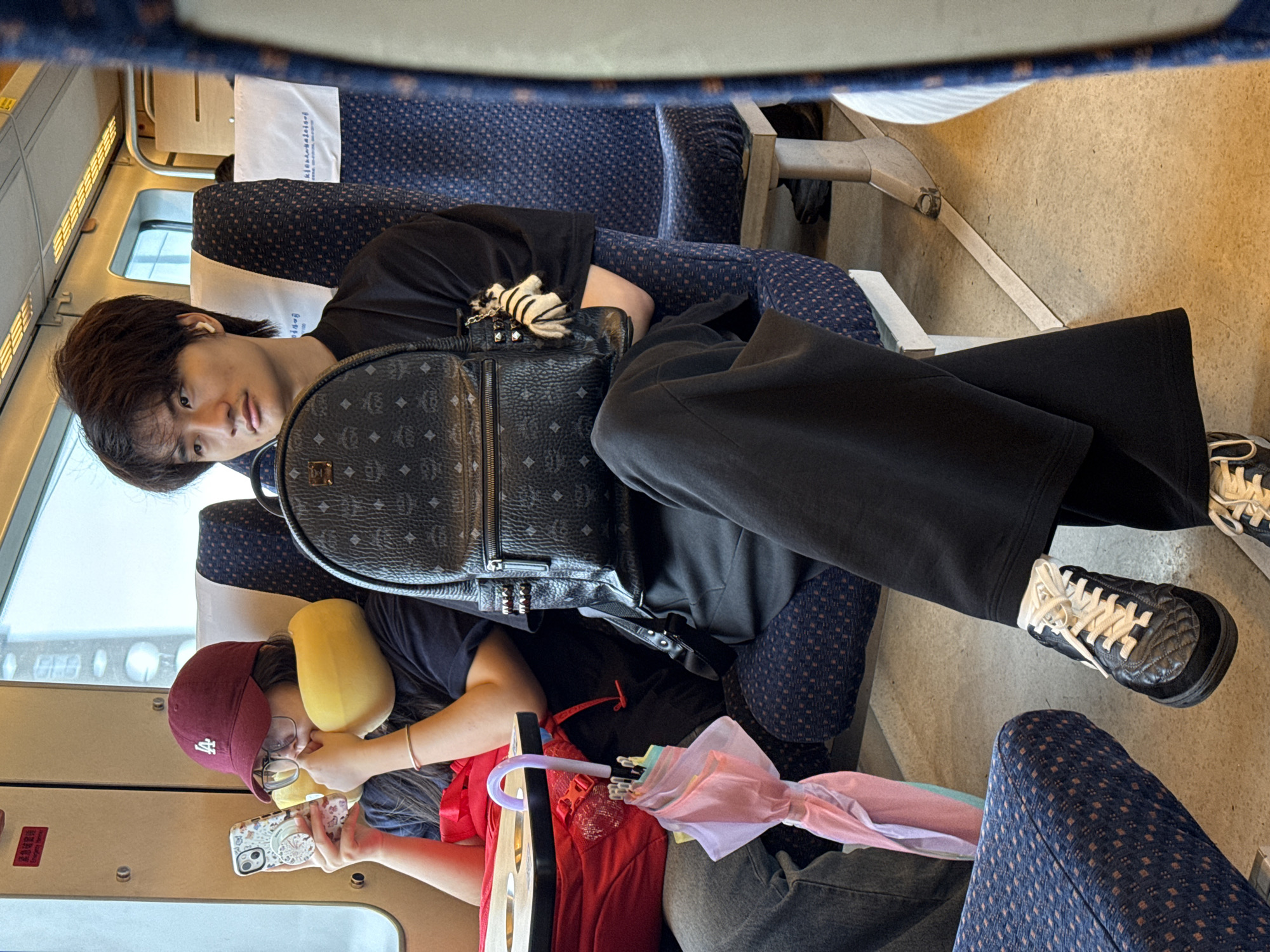 On a train, an individual with dark hair and a black t-shirt sits next to a window and wears black pants. An MCM backpack rests on their lap. Beside them, another person, wearing a maroon baseball cap and a yellow neck pillow, looks at a mobile phone. A partly open multi-colored umbrella rests beside them. The train seats are upholstered in a blue patterned fabric. The location is the city of Baiyun District, China.