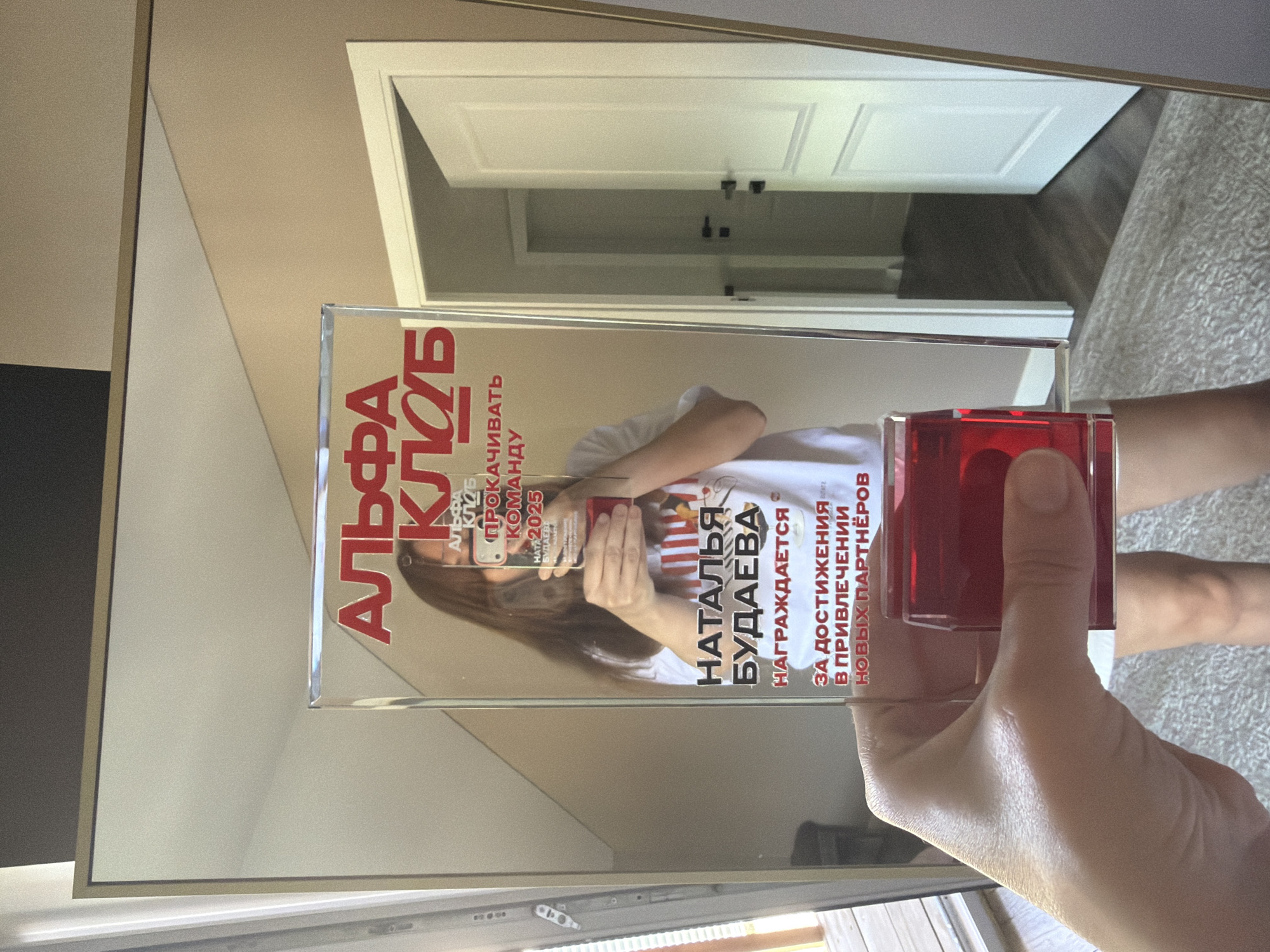 A person is holding a clear award with red lettering in front of a mirror. The award is rectangular and appears to be made of glass or acrylic. The lettering on the award reads "АЛЬФА КЛУБ" at the top, followed by "ПРОКАЧИВАТЬ КОМАНДУ 2025". Below this, it says "НАТАЛЬЯ БУДАЕВА НАГРАЖДАЕТСЯ ЗА ДОСТИЖЕНИЯ В ПРИВЛЕЧЕНИИ НОВЫХ ПАРТНЁРОВ". A red, liquid-filled base is attached to the bottom of the award. The person's hand is visible, holding the award. The mirror reflects a room with a white door and a rug. The overall scene suggests an indoor environment, possibly a home or office. The text on the award is in Russian. There is no indication of the specific location within the city of Malinovka, Russia.