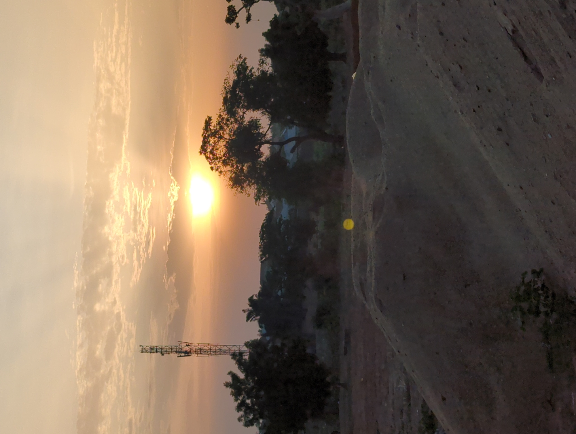 The image depicts a sunset scene in what appears to be a rural area of Narayi, Nigeria. The sky is filled with orange and yellow hues due to the setting sun, which is partially obscured by clouds. Silhouetted against the sky are trees and a cell tower. In the foreground, there is a large rock formation with textured, gray surfaces. Vegetation grows near the base of the rock. The overall environment suggests a location on the outskirts of the city.