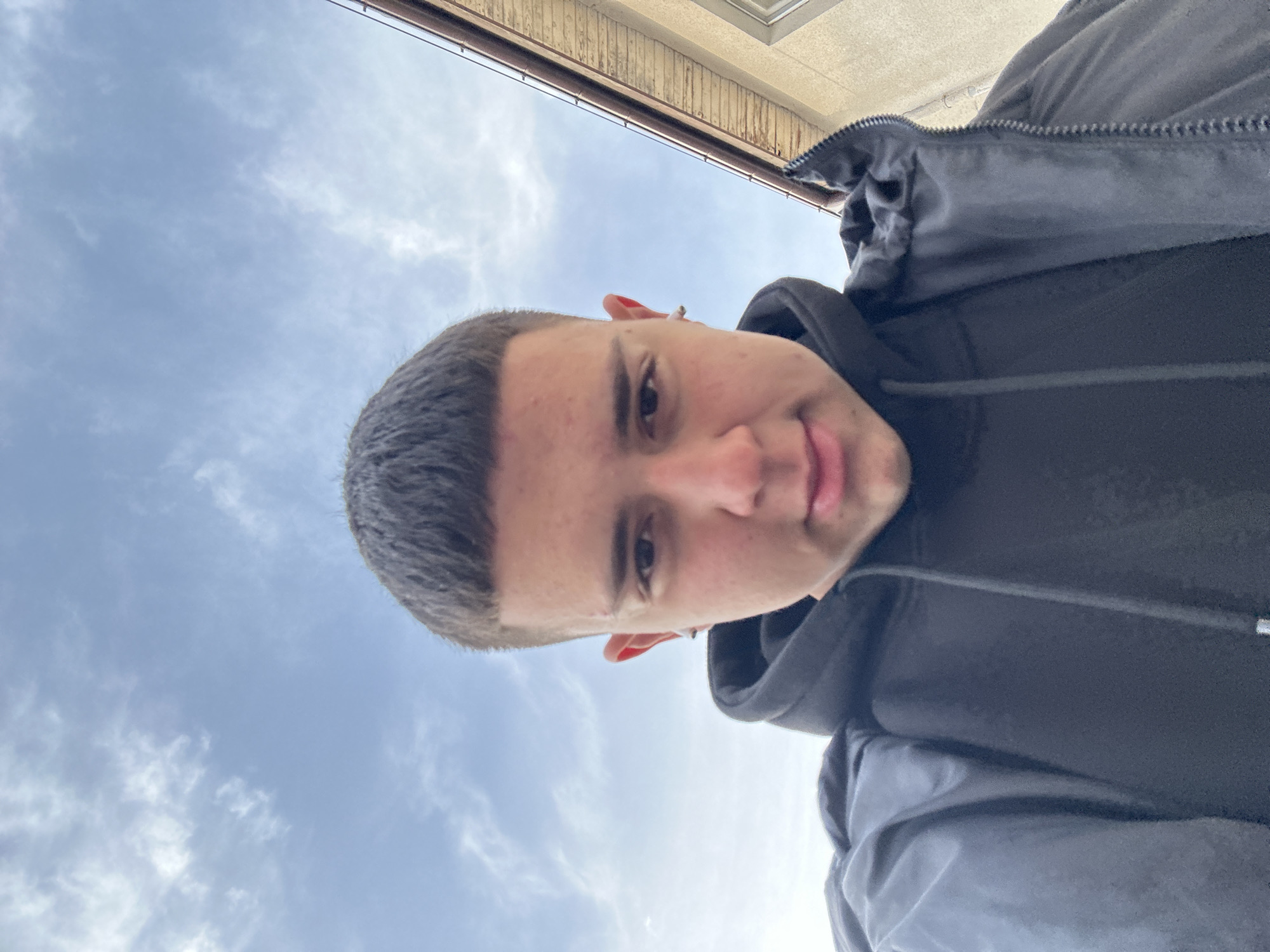 A low-angle self-portrait of a young man. The subject has short, dark hair and is wearing a black hooded sweatshirt and a dark-colored jacket. The background shows a cloudy sky and the top of a building with brown wooden eaves. The location is assumed to be Zlatograd, Bulgaria.