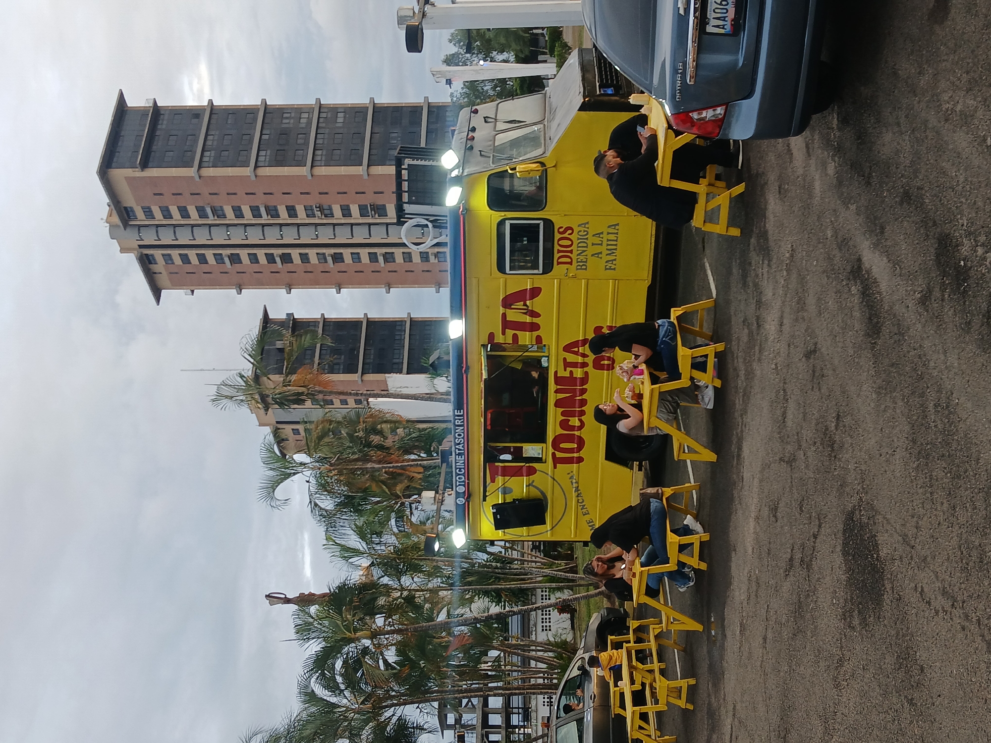 The image depicts an outdoor dining scene at dusk in Ciudad Guayana, Venezuela, centered around a yellow food truck. The food truck, labeled "TO CINETA" in large red letters and "DIOS BENDIGA A LA FAMILIA" (God Bless the Family), is brightly illuminated by multiple roof-mounted lights and features a speaker and a smiley face graphic. A sign on the top left of the truck reads "OTO CINE TASON RIE".
In front of the food truck, five yellow wooden picnic tables with attached benches are arranged on an asphalt surface. Patrons occupy four of these tables. On the far left, a woman with dark hair and a young boy are seated at a table. At the next table, two women with dark hair are seated, one looking at her phone and the other smiling towards the right. To the right, two men are seated at a table; one is looking at his phone while the other leans towards him. Another person is partially visible inside the food truck, behind a serving window.
Two vehicles are visible: a dark grey/brown sedan on the far left with a person in the driver's seat, and a dark blue Chevrolet Optra 1.8 sedan on the far right, partially visible with its rear and Venezuelan license plate "AA063P" facing the viewer.
The background features numerous tall palm trees and two modern, multi-story buildings with a mix of light brown/beige and dark glass facades. The sky is overcast and light grey, indicating evening or late afternoon conditions. A white pole with a light fixture is visible on the far right. The ground is dark grey asphalt with signs of wear.