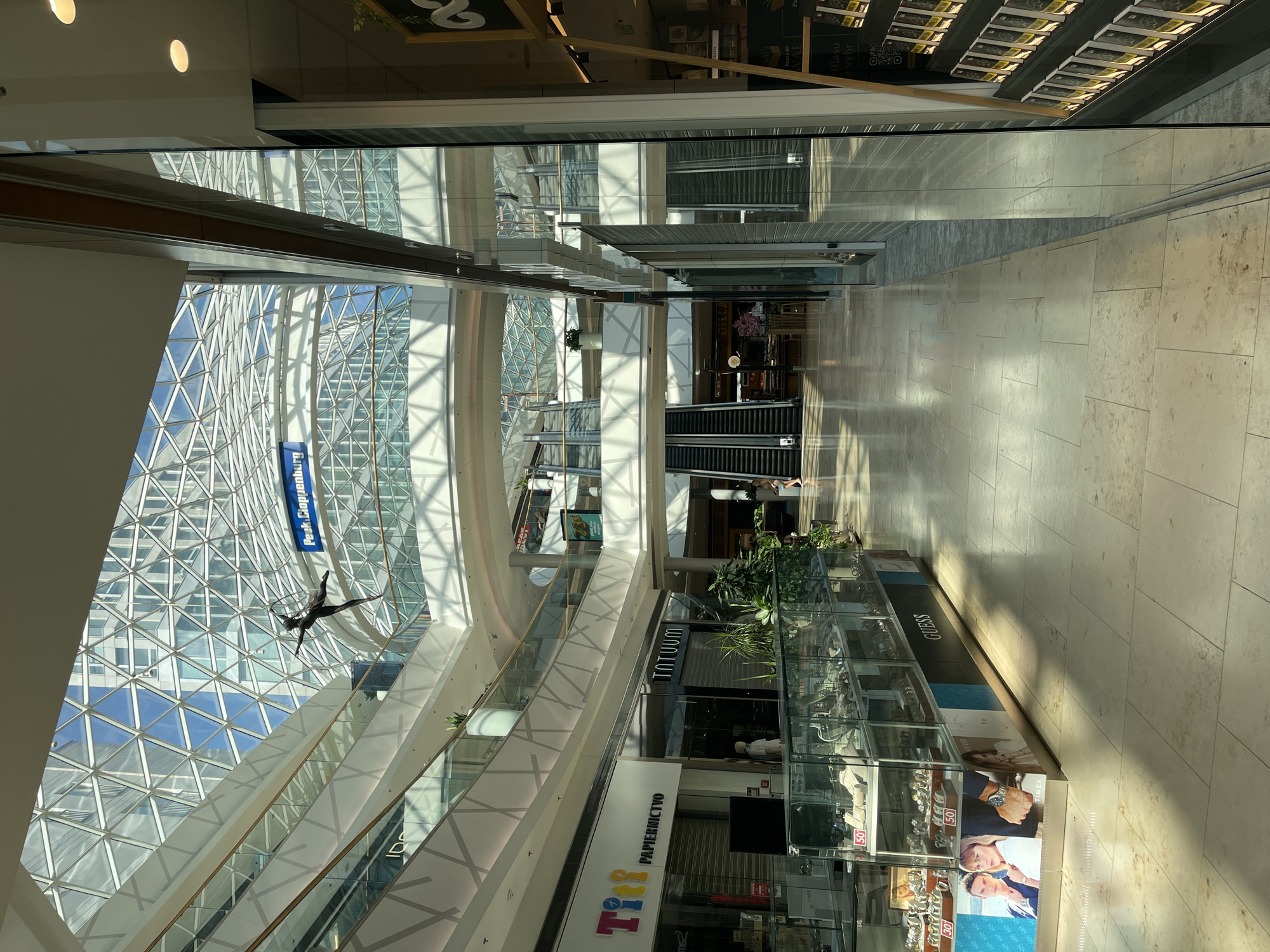The image shows the interior of a modern shopping mall with a large, glass-paneled, geometric-patterned ceiling that allows natural light to flood in. Suspended from the ceiling by thin wires is a metallic sculpture of an acrobatic figure in mid-air. The mall features multiple levels with white-paneled balconies and escalators visible in the background. On the ground floor, shops line the wide, tiled walkway. To the left, a jewelry store display case showcases watches and other accessories. A "Tiff" stationery store and a "Tatuum" clothing store are also visible. Further down the walkway, escalators lead to the upper levels. A person is seen walking on the right side of the walkway. The mall appears to be the Eurovea shopping center in Bratislava, Slovakia, given the signage for "Pestloppensburg," which is an older name for the area and likely a reference in the mall's context.