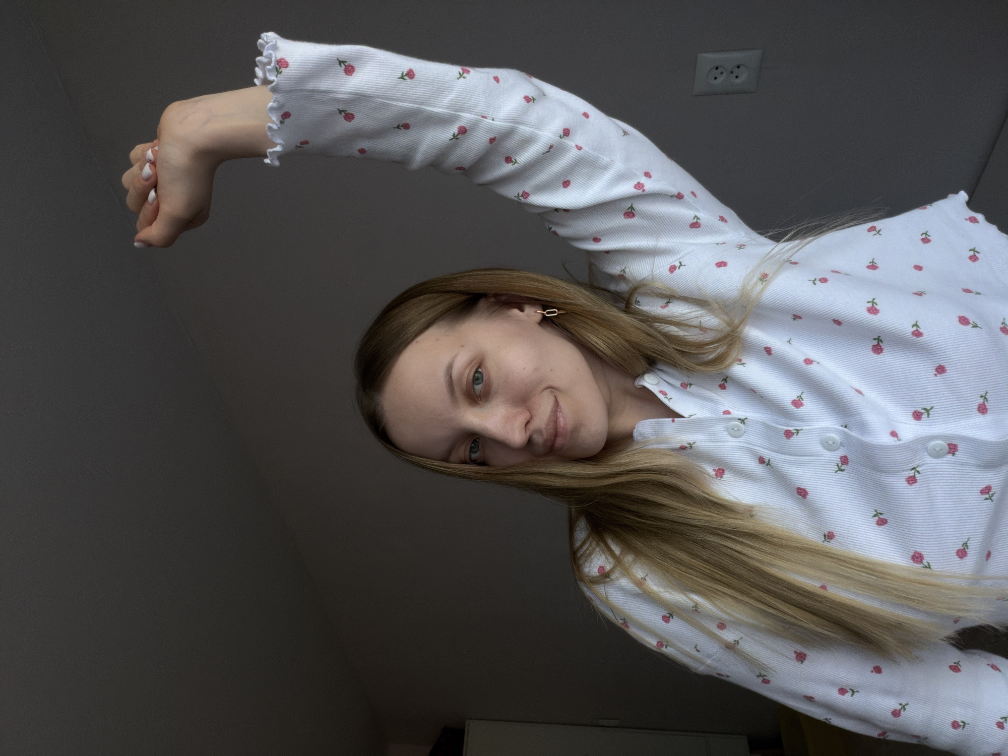A young woman with long blonde hair, wearing a white long-sleeved top with a pattern of small pink flowers, is photographed from a slightly low angle. The shirt has a ruffled trim at the end of the sleeves and buttons down the front. She is extending her arm above her head, with her hand appearing near the top left of the frame. Her nails are painted white. She is looking downward with a slight smile. The background is a gradient of grey to darker grey, suggesting a dimly lit room. An electrical outlet with two sockets is visible on the wall near the upper center of the frame. The location details are not apparent from the provided image. Therefore, specifying the exact location as Reshetnikovo, Russia, cannot be confirmed based on the visual information alone.