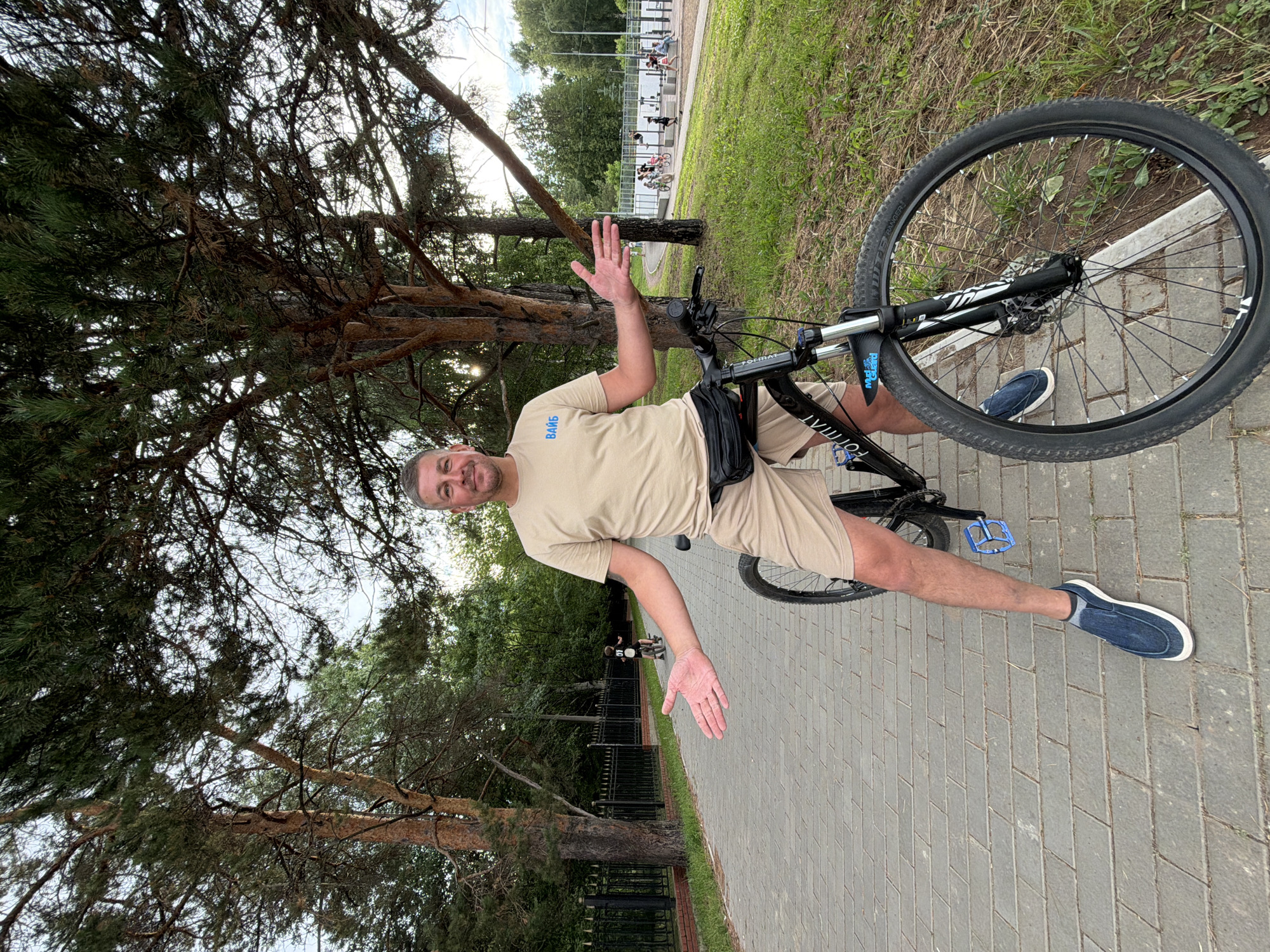 A man in a tan t-shirt and shorts stands next to a black bicycle on a paved walkway in a park in Chaikovsky, Russia. The man has his right arm extended, palm open, as if gesturing. The bicycle is positioned in front of him, and its front wheel is in the foreground. Behind the man, a child on a bicycle is visible further down the path, and in the background, there is a playground with people. The environment is characterized by trees, grass, and a fence. The sky is overcast, suggesting it may be late afternoon or early evening. The paved walkway is made of interlocking pavers, and the grass is green and well-maintained. The trees are tall and leafy, with some pine trees also present. The fence appears to be made of wrought iron.