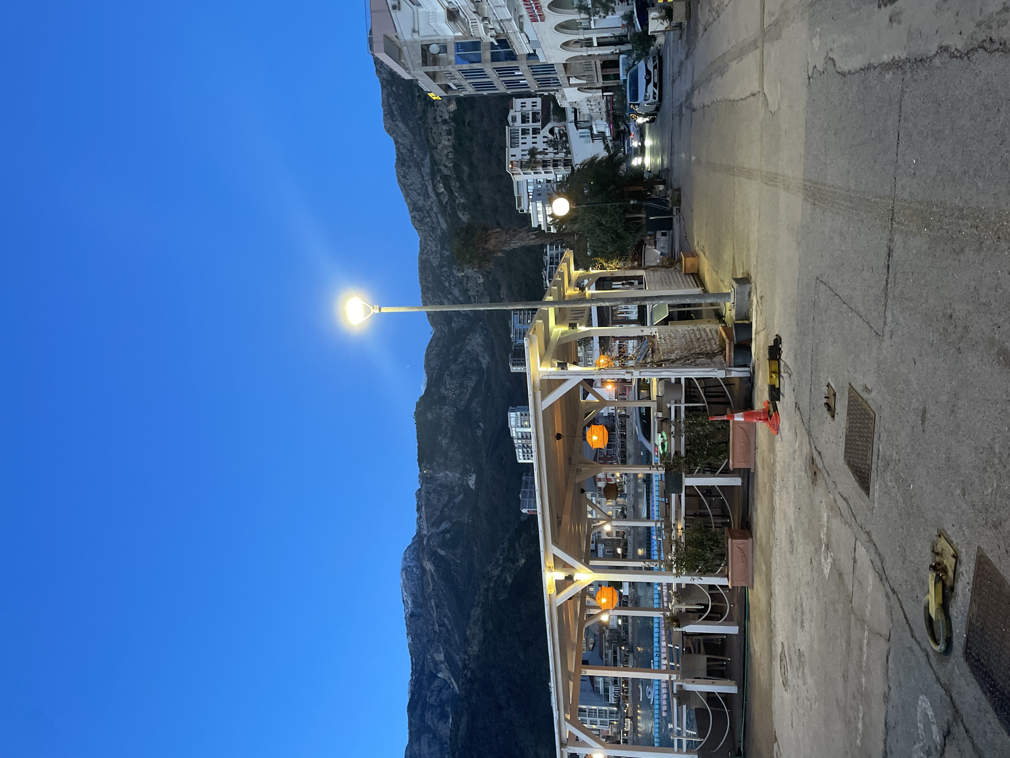 The scene depicts a coastal urban environment at dusk in Budva, Montenegro. A deep blue twilight sky covers a landscape dominated by large, rugged mountains in the background, showing traces of lighter rock or sparse vegetation near their peaks.
In the mid-ground, an illuminated, open-air restaurant or cafe structure, characterized by a white wooden framework and a flat roof, occupies the left side of the image. Multiple spherical, orange pendant lights hang within the structure, illuminating wicker-style chairs and tables. Planters with green foliage are arranged along its perimeter, and a rope barrier separates it from a body of water. Several small boats or floating platforms are visible on the water, reflecting distant lights.
A paved road, exhibiting tire tracks and surface cracks, runs through the center and right of the image, curving slightly to the right towards the background. A tall, modern streetlight with a bright, glowing luminaire stands prominently near the center-right, casting light onto the road and adjacent structures. Further down the road, another streetlight is visible, and several vehicles with illuminated headlights are present, including a white sedan parked on the right side.
Along the right side of the road, multi-story buildings of varying architectural styles are visible, including one with modern glass facades and another with arched ground-level entrances displaying commercial signage such as "ZEE" and "MANANA." A tall palm tree is situated behind the prominent streetlight.
The immediate foreground features a concrete or asphalt surface with metal manhole covers and a yellow and black ground anchor. An orange traffic cone lies on its side near the restaurant structure. No individuals are visibly present or engaged in activity within the depicted area.