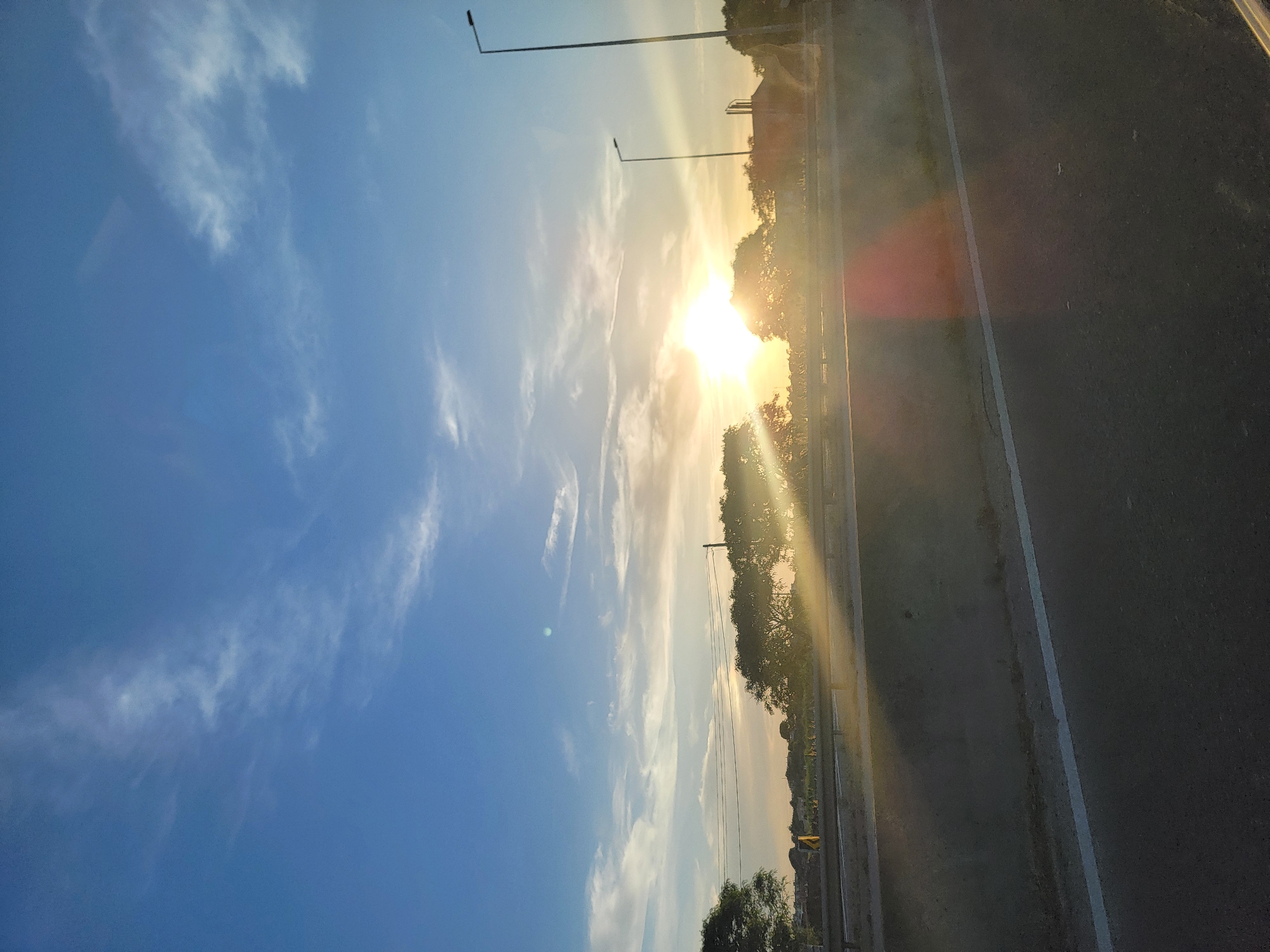The image depicts a sunset over a highway in Faro, Portugal. The sun is low on the horizon, casting long rays of light across the sky and the landscape. The sky is a deep blue, with scattered white clouds. Trees line the side of the highway, their branches silhouetted against the bright sun. A concrete barrier separates the highway lanes, and streetlights are visible in the distance. The road surface is dark asphalt, with a white line marking the edge. The overall impression is one of a peaceful, serene evening.