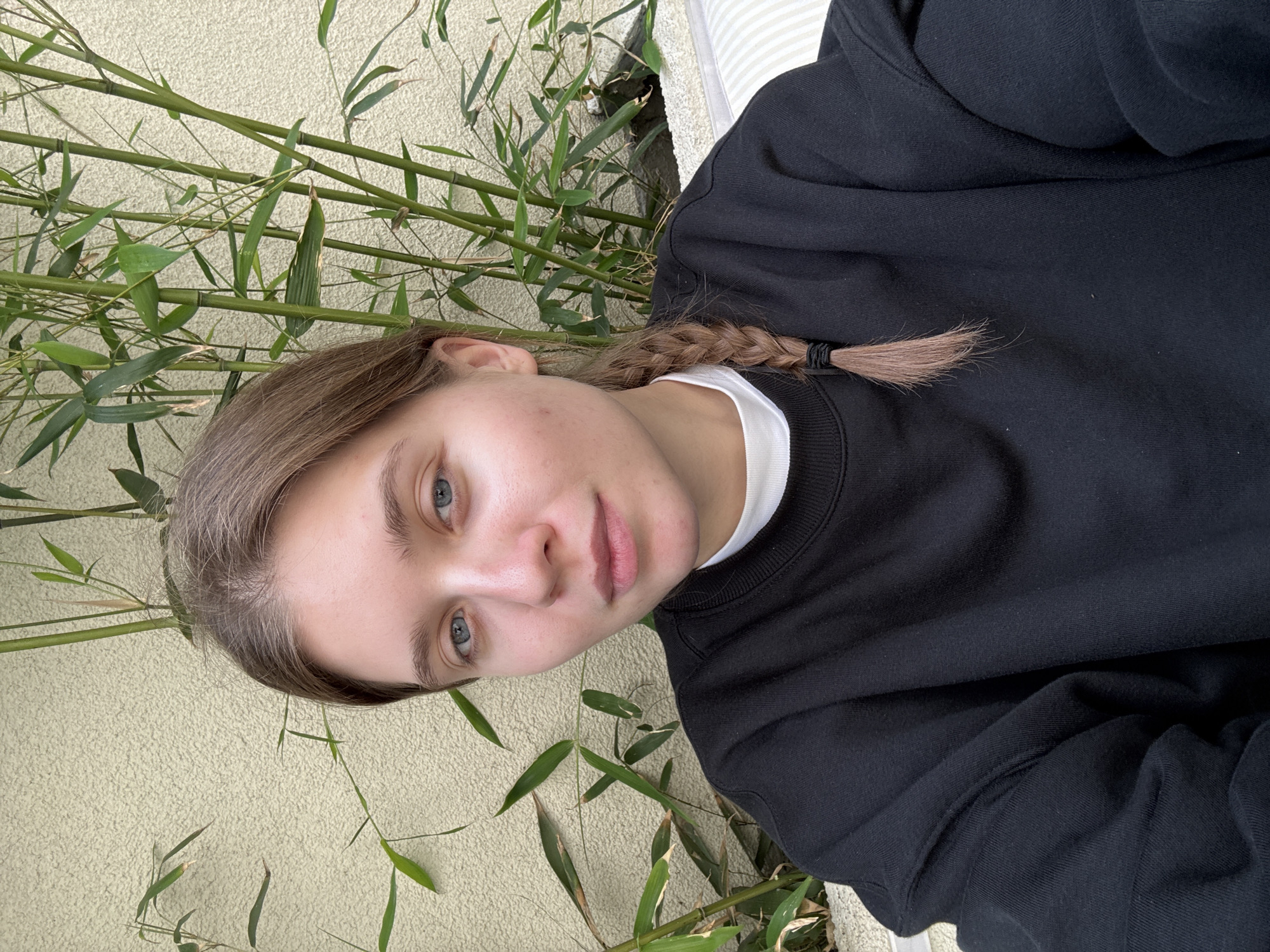A woman with light brown hair braided at the end is wearing a black sweatshirt with a white collar peeking through. She has fair skin and blue eyes. The background includes a textured beige wall and green bamboo stalks with leaves. It is impossible to determine the location in Belgrade, Serbia, or any actions or interactions without more information.