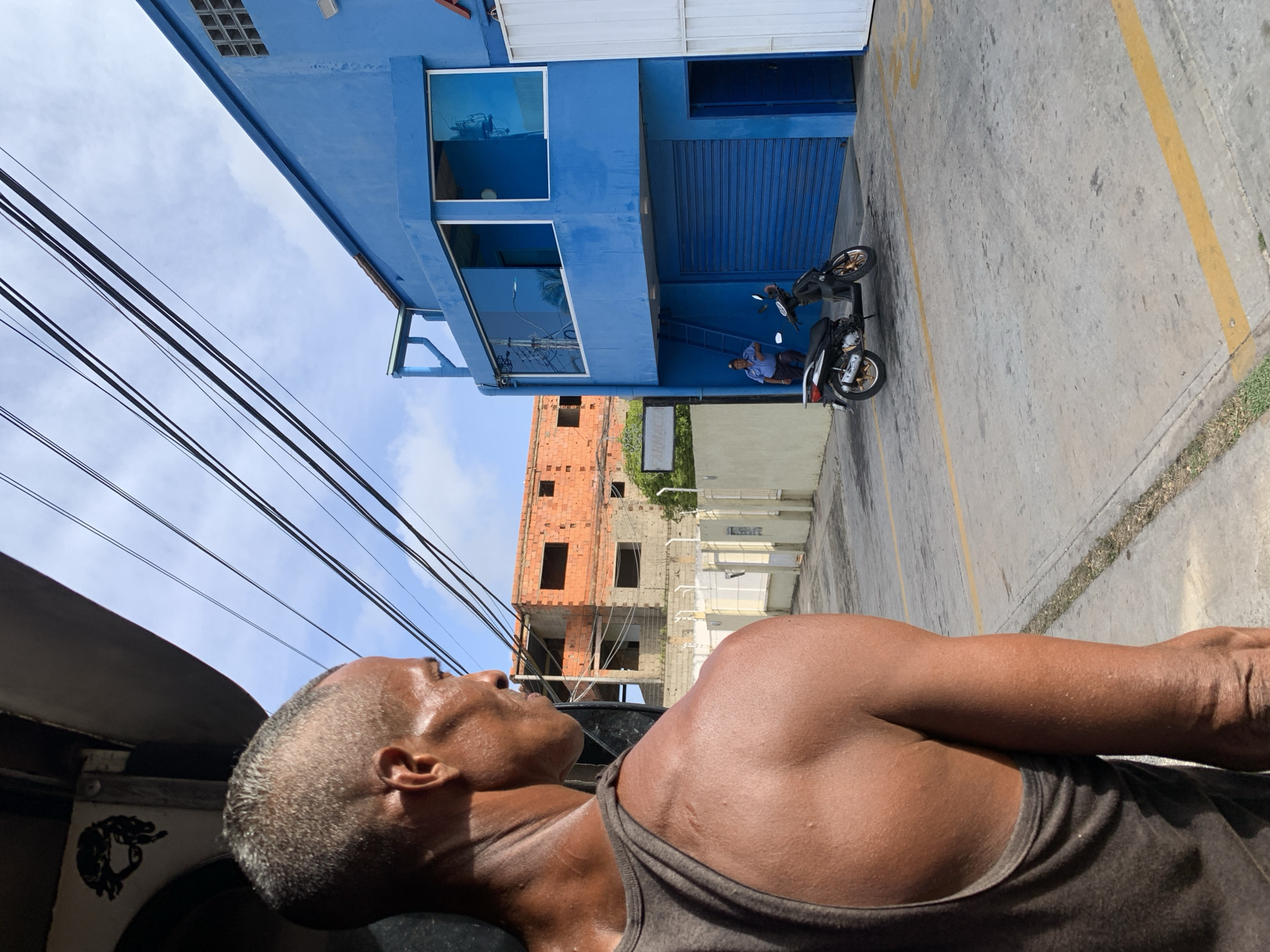 The image captures a vibrant street scene, looking upwards from a low vantage point, possibly from inside a vehicle. In the immediate foreground, to the right, a man with dark skin and short, possibly graying hair, is visible from the chest up. He wears a dark tank top, and his skin appears shiny, suggesting warmth or activity. He looks upwards and to the left, observing the scene. A black graphic, possibly an animal head, is visible on the vehicle he seems to be in or beside, in the bottom left corner.
The main focus in the midground is a striking two-story building painted a bright royal blue. It features large reflective windows on the upper floor, showing reflections of the sky and surrounding elements. The ground floor has a blue roll-down garage door. Adjacent to this building, a man in a light blue collared shirt and dark pants stands beside a black motorcycle with gold-colored wheels, suggesting a daily routine.
Behind the blue building, an unfinished reddish-orange brick structure with multiple empty window openings stands, hinting at ongoing construction or an abandoned project. Further back, other light-colored concrete buildings are partially visible. Overhead, several thick black utility wires crisscross against a clear, bright blue sky with a few white clouds. The concrete ground below is marked with yellow lines, possibly for parking, and some patches of grass or weeds line the edges. Yellow numbers, partially obscured, are visible on the concrete, appearing to be "400".
The bright sunlight and clear sky indicate it is daytime, likely a warm day. The scene depicts a slice of daily life in what appears to be an urban or semi-urban environment, consistent with a location like La Asuncion, Venezuela.