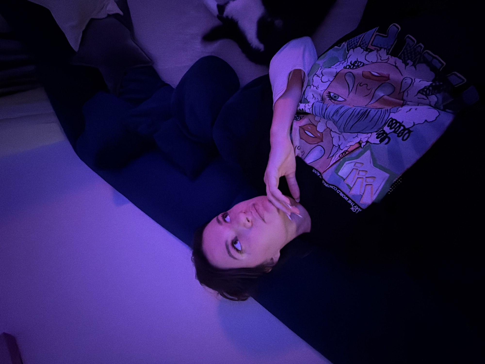 A woman is lying on a dark, possibly black, surface. She's wearing dark clothing, possibly black sweatpants and a t-shirt. The t-shirt has a white collar and a graphic print with cartoon-style faces in shades of white, light blue, and pink. She has brown hair and fair skin and is touching her face with her right hand. The background appears to be a room with a white or light-colored wall on the left, lit with a strong purple light. The rest of the room is dark. At the top of the frame, out of focus, is a white object, possibly a pillow, and a black and white object, possibly a cat or blanket. There is no information to indicate this photo was taken in Belgrade, Serbia.