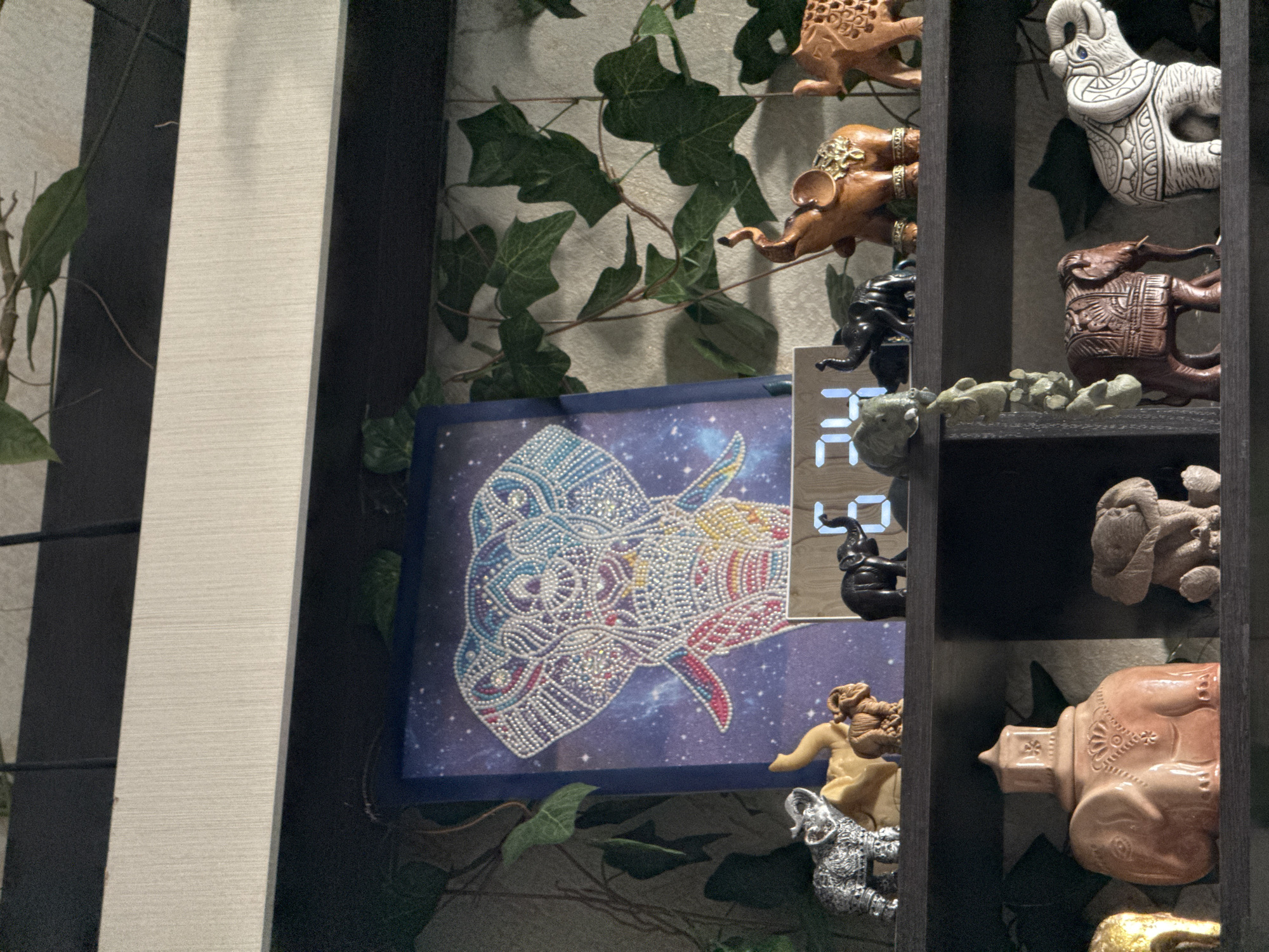 The image shows a shelf decorated with elephant figurines. There are several shelves visible, each containing multiple figurines of varying sizes and colors. The figurines appear to be made of different materials, including ceramic, wood, and possibly metal. Behind the shelf, there's a backdrop of artificial greenery with climbing vines. A framed artwork featuring an elephant's head rendered in a pointillist style with cosmic background is also visible. A digital clock is partially visible, showing numbers in a blocky, LED style. The setting appears to be indoors, likely a private residence. It is not possible to determine if this image was taken in Ново-Никольское, Russia without additional context or information.