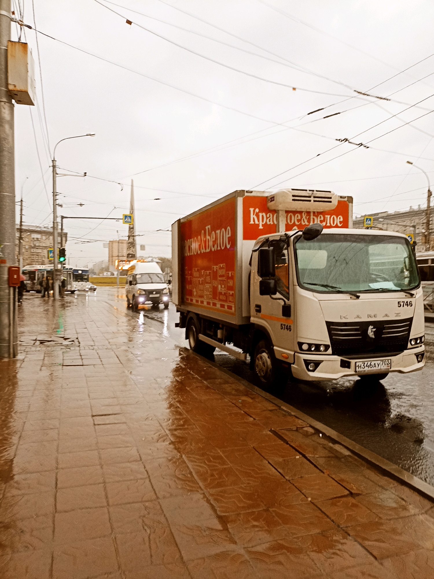 A wet, overcast day is depicted in this urban scene, possibly in Russia. A white Kamaz truck with an orange box body, advertising "Красное & Белое" (Krasnoe & Beloe), is the primary subject. The truck has the license plate H346AY 702. To the left of the Kamaz truck, a white van is visible. Further down the street, a bus with the number 5746 can be seen, along with a traffic light that is currently green. The sidewalk is paved with brown tiles and is reflecting the wet conditions. Overhead, numerous wires crisscross the sky, suggesting an urban environment with trolleybus or power lines. In the background, buildings and a tall, obelisk-like structure are present. The overall atmosphere is damp and subdued due to the weather.