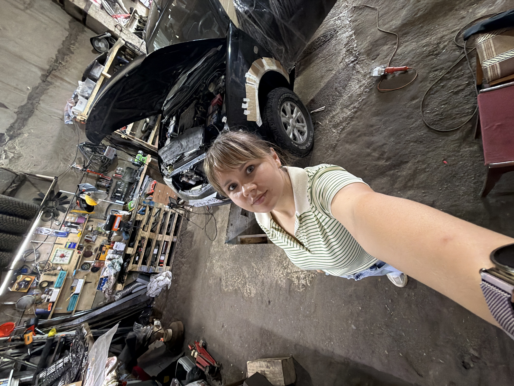The image depicts a cluttered garage with a black car with its hood open. A woman in a striped shirt is taking a selfie, her arm extended towards the camera. Various tools and car parts are scattered around the garage, including shelves filled with items, car tires stacked in the background, and a red car jack on the floor. The overall environment suggests an active workshop or repair space. The image is tagged as being located in Chaikovsky, Russia.