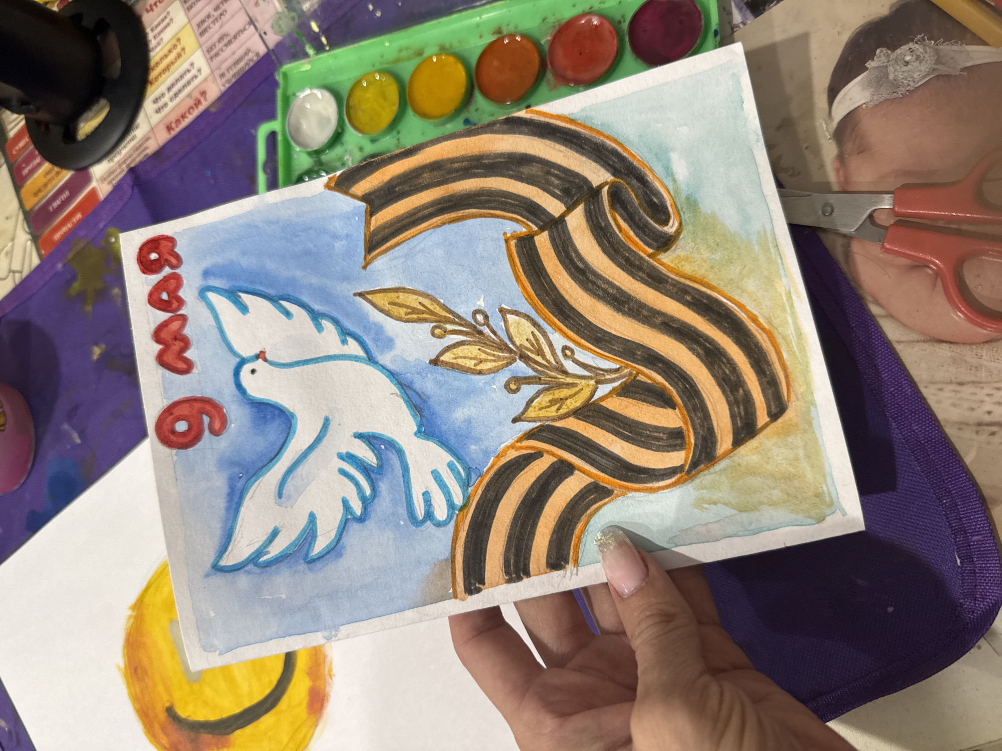 The image shows a hand holding a watercolor painting. The painting features a white dove in a blue sky with a St. George ribbon and a branch with golden leaves. The red inscription "9 мая" (May 9th) is visible.
The scene includes art supplies, such as a watercolor palette with paint in different colors and a sheet with Russian grammatical cases, indicating an art or craft activity.
The image's location is not mentioned in the text, and the city of Rusanovo, Russia is not discernible from the image.