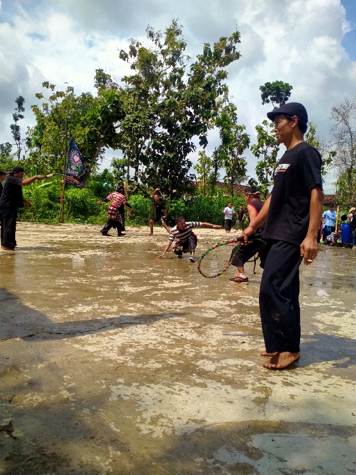 The image shows a group of men in a clearing in a wooded area in Kecamatan Kedawung, Indonesia (latitude: -7.518, longitude: 111.029). The men are gathered in a muddy clearing. Some of the men are practicing a martial art, while others are standing around watching. One man in a black t-shirt and black pants is holding a long rope and standing near the edge of the clearing. He is watching the men practice. There is a large tree with leafy branches in the background. There is also a flag hanging from a pole in the background. The ground is covered with mud and the sky is mostly clear.