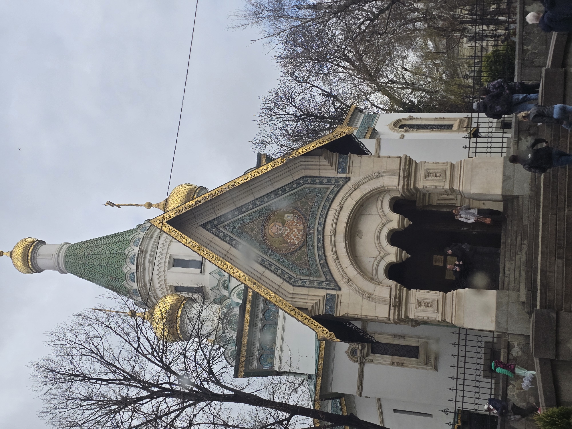The image depicts the Church of St. Nicholas the Miracle-Maker in Sofia, Bulgaria. The church has a green-tiled roof and gold domes. The front facade is decorated with mosaics, and the entrance is accessed by stairs. People are seen standing on the stairs and entering the church. A wrought iron fence is visible on the right side of the image. Bare trees frame the church against an overcast sky.