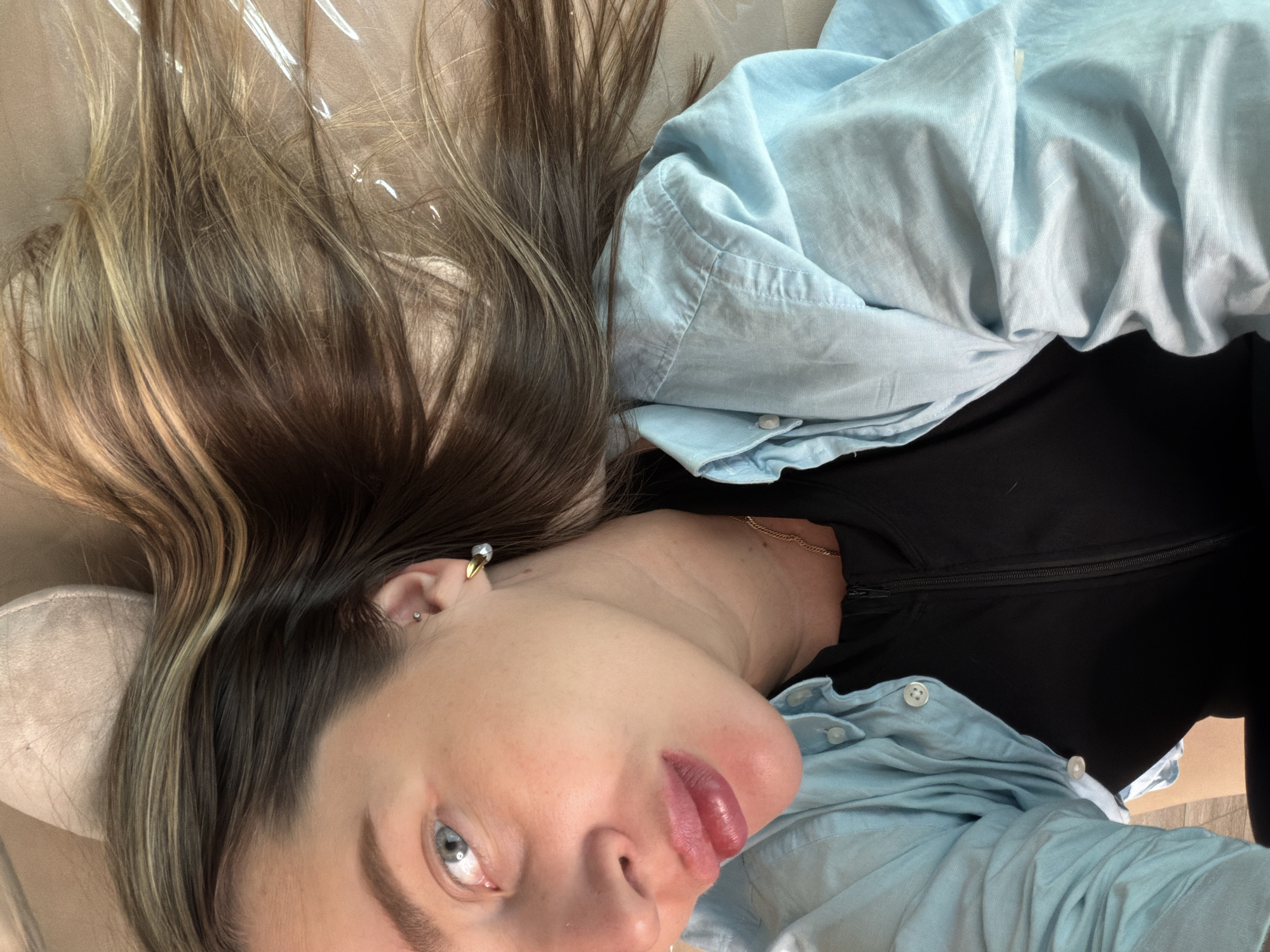 A young adult woman lies supine on a surface covered with a translucent material, likely plastic. She has light skin, blue eyes, and dark brown hair with blonde highlights. She wears a light blue button-down shirt over a black top. A gold chain necklace is visible on her neck, and multiple earrings adorn her left ear. The background appears to be a neutral tan color. The image provides no information to suggest that it was taken in the city of Moscow, Russia.