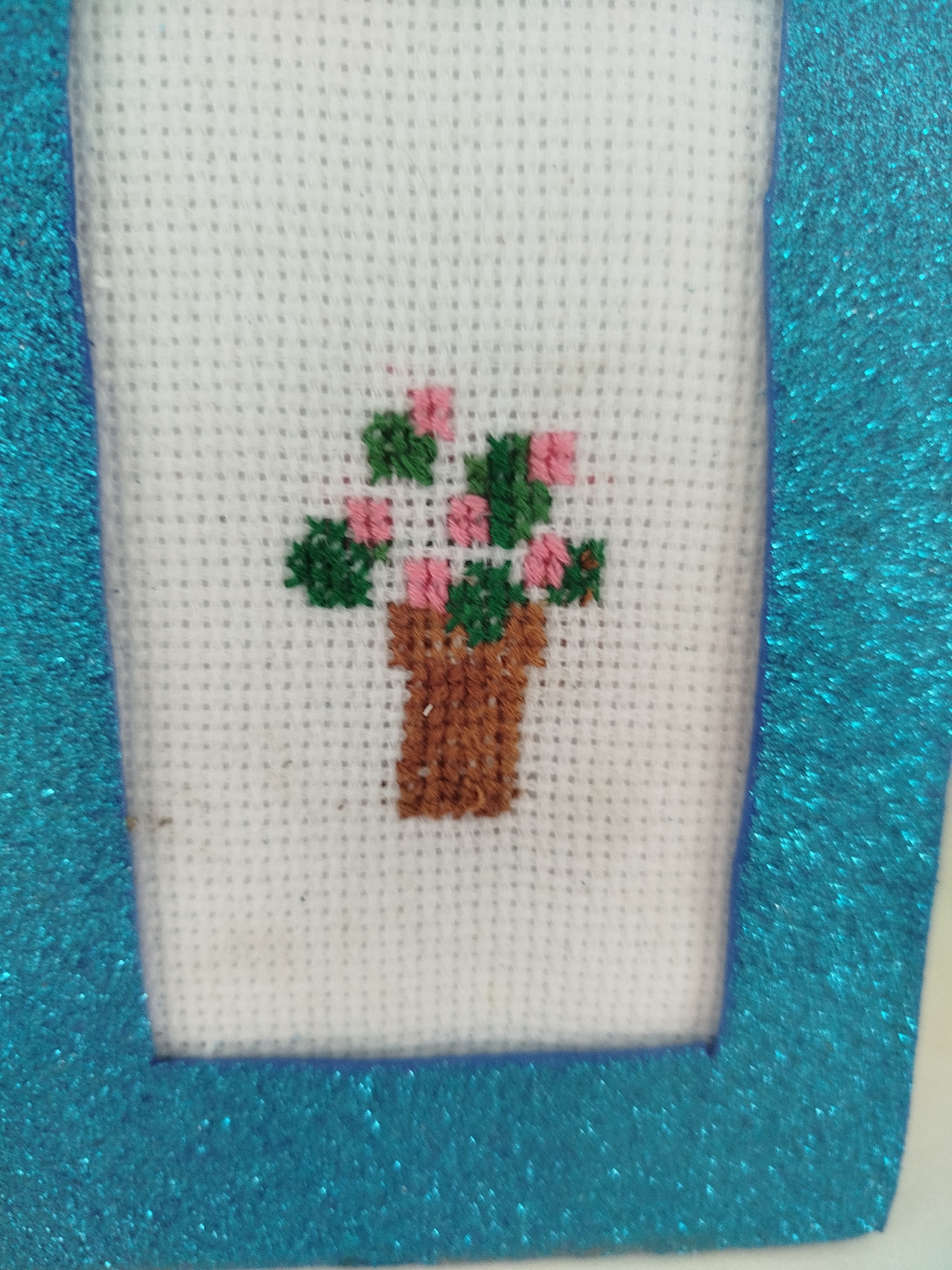 The image shows a close-up of a cross-stitch embroidery featuring a small potted plant with pink flowers and green leaves. The embroidery is on white Aida fabric and is framed by a textured, glittery blue material. The background is out of focus. The specific location in Крывасельскі сельскі Савет, Belarus is not discernible from the image.
