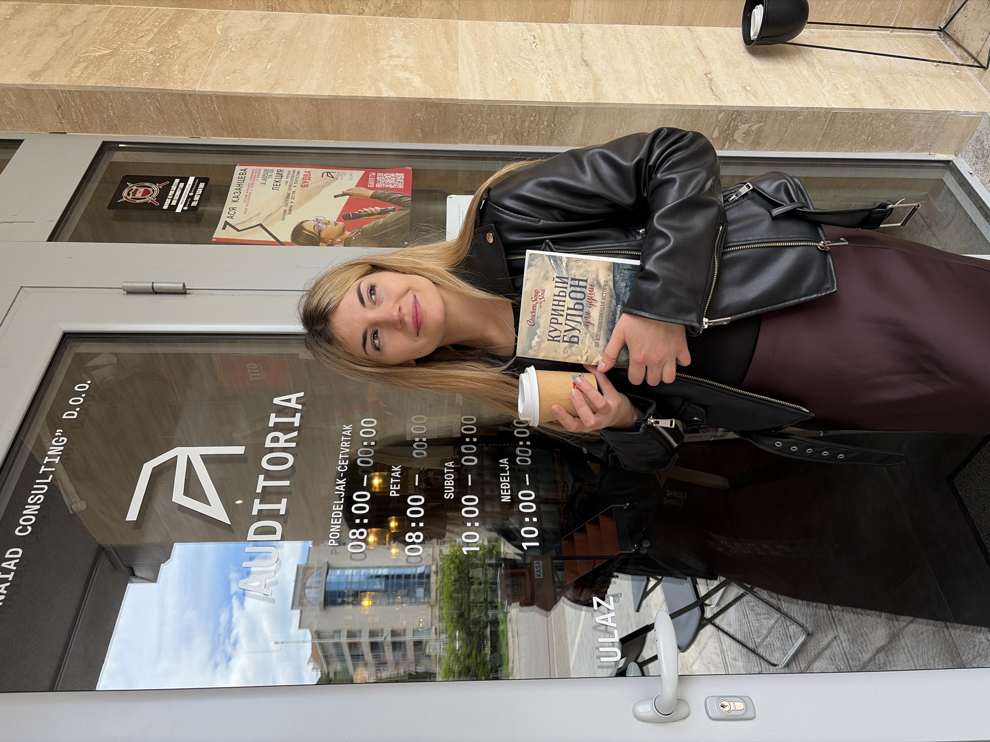 A young woman stands in front of a glass door, holding a book and a coffee cup. The book is titled "Chicken Soup for the Soul" in Russian ("Куриный бульон для души"). The door belongs to "AUDITORIA", a consulting firm. Operating hours are listed: Monday-Thursday 8:00-00:00, Friday 8:00-00:00, Saturday 10:00-00:00, Sunday 10:00-00:00. A poster advertises a lecture by Asya Kazantseva on April 3rd at 19:00 in Budva. The address is Tito, Budva, Montenegro.