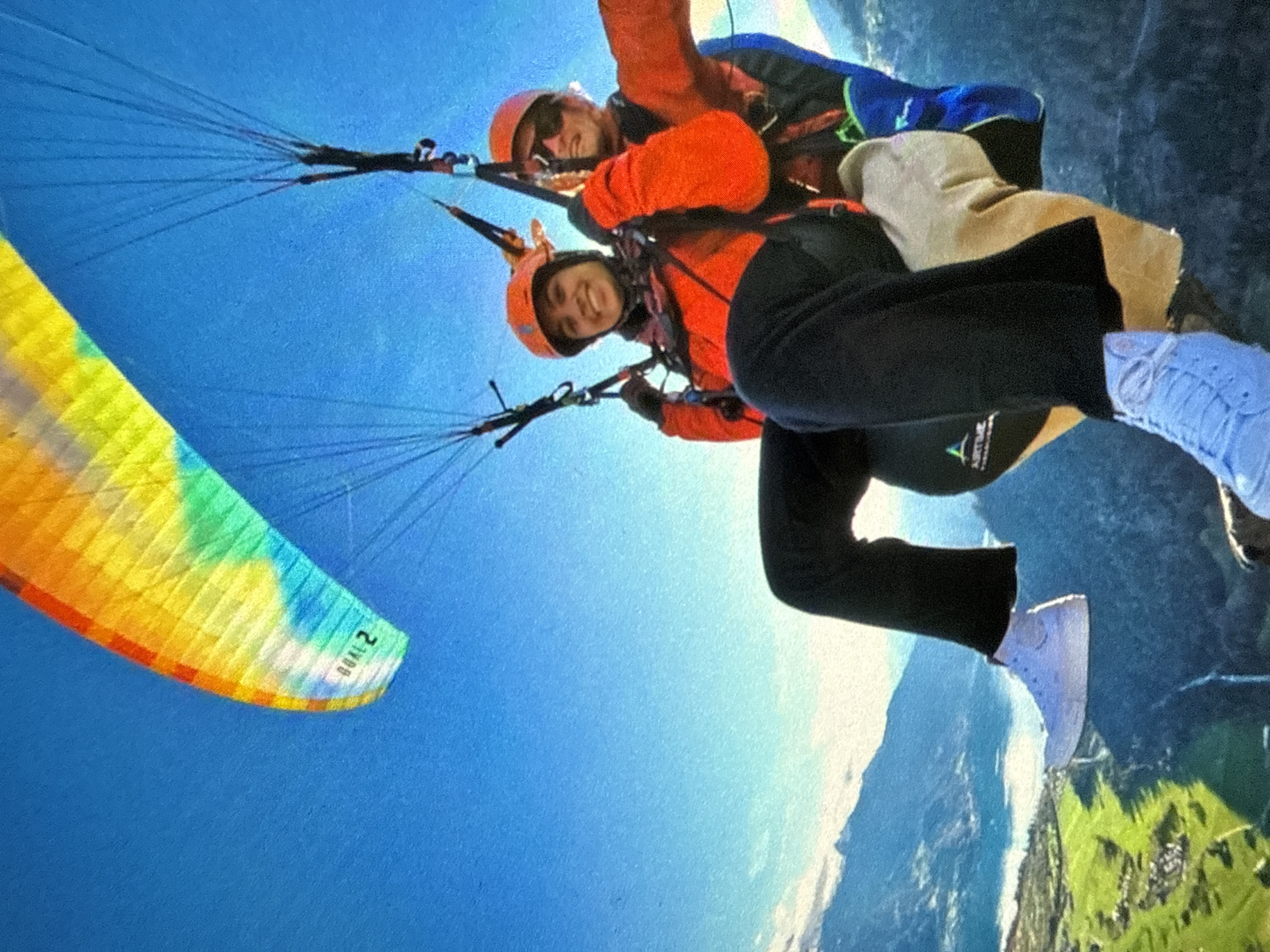 Two people are tandem paragliding against a mostly clear blue sky. They are wearing helmets and orange jackets. The person in the front has black pants and white sneakers. The paragliding wing is multi-colored (yellow, green, blue) and has the word "GOAL 2" on its edge. Below them, a mountainous landscape with green patches is visible. The location is Unknown, Unknown.