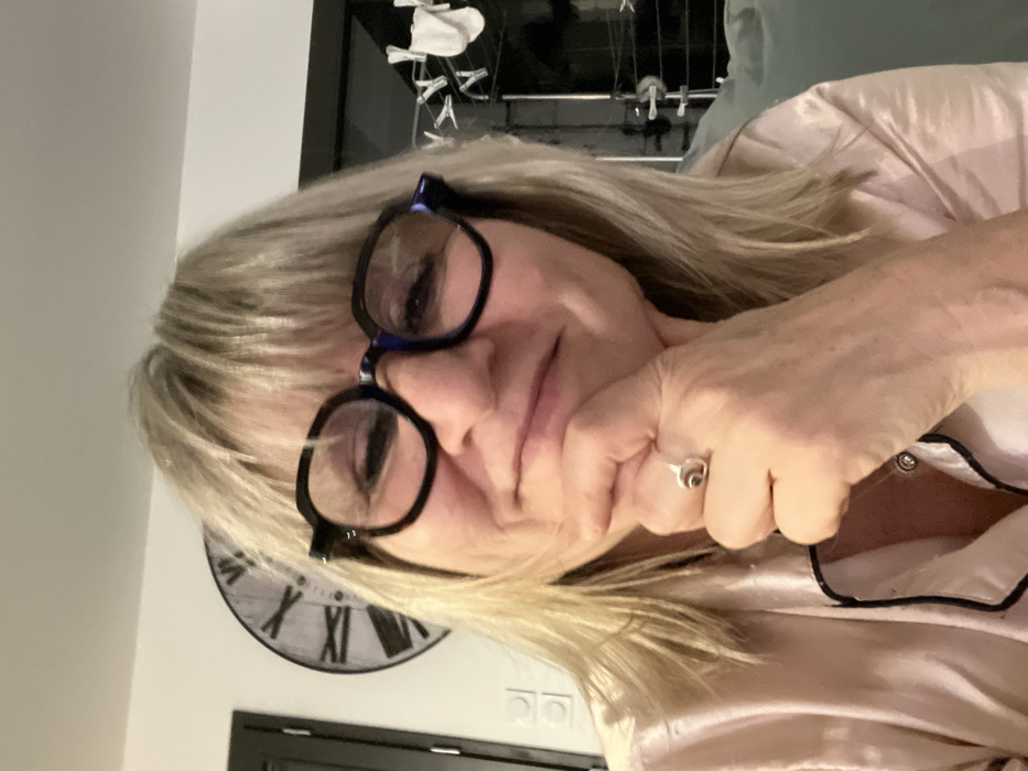 A woman with blonde hair and black-rimmed glasses rests her chin on her hand. She's wearing a pink satin pajama top with black trim. Her ring is visible on her hand. In the background, a black-and-white clock with Roman numerals is visible. Behind her, a glimpse of a rack with clothes hanging on it.
