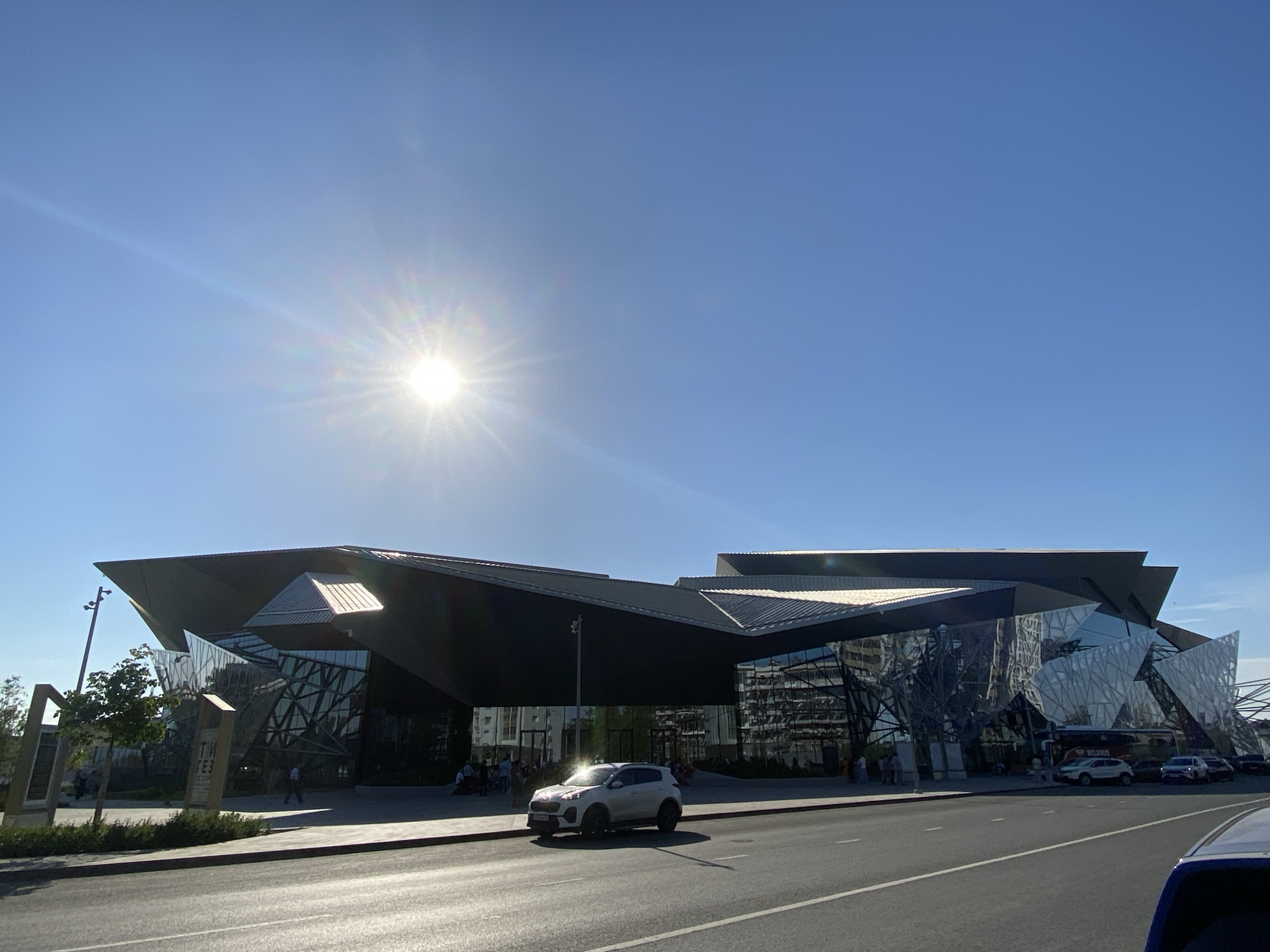 The image depicts the exterior of the Kazan Family Center ("Kazan" National Cultural Center) in Kazan, Russia, on a bright, sunny day. The building is a modern structure with angular, geometric shapes and reflective glass surfaces. A white Kia Sportage SUV is parked in front of the building. Several people can be seen walking near the entrance. A road is in the foreground, with a blue car partially visible in the lower right corner.