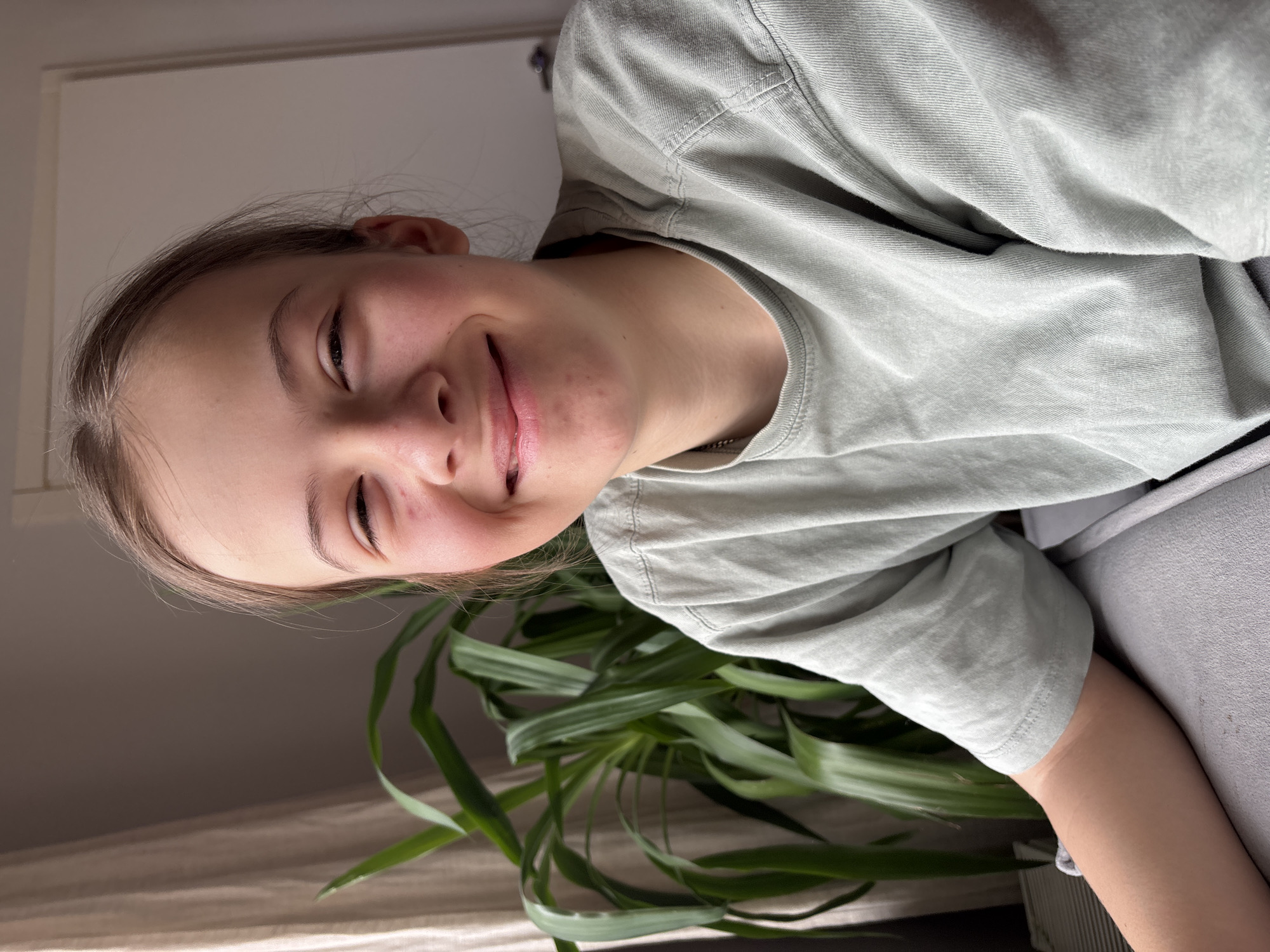 A young person with fair skin, light brown hair pulled back, and wearing a light green t-shirt is smiling slightly with eyes closed. They are indoors with a large leafy plant visible in the lower portion of the frame. A portion of what appears to be a light-colored curtain and a white wall are also visible. There is no information provided in the image to indicate this is Chaikovsky, Russia.