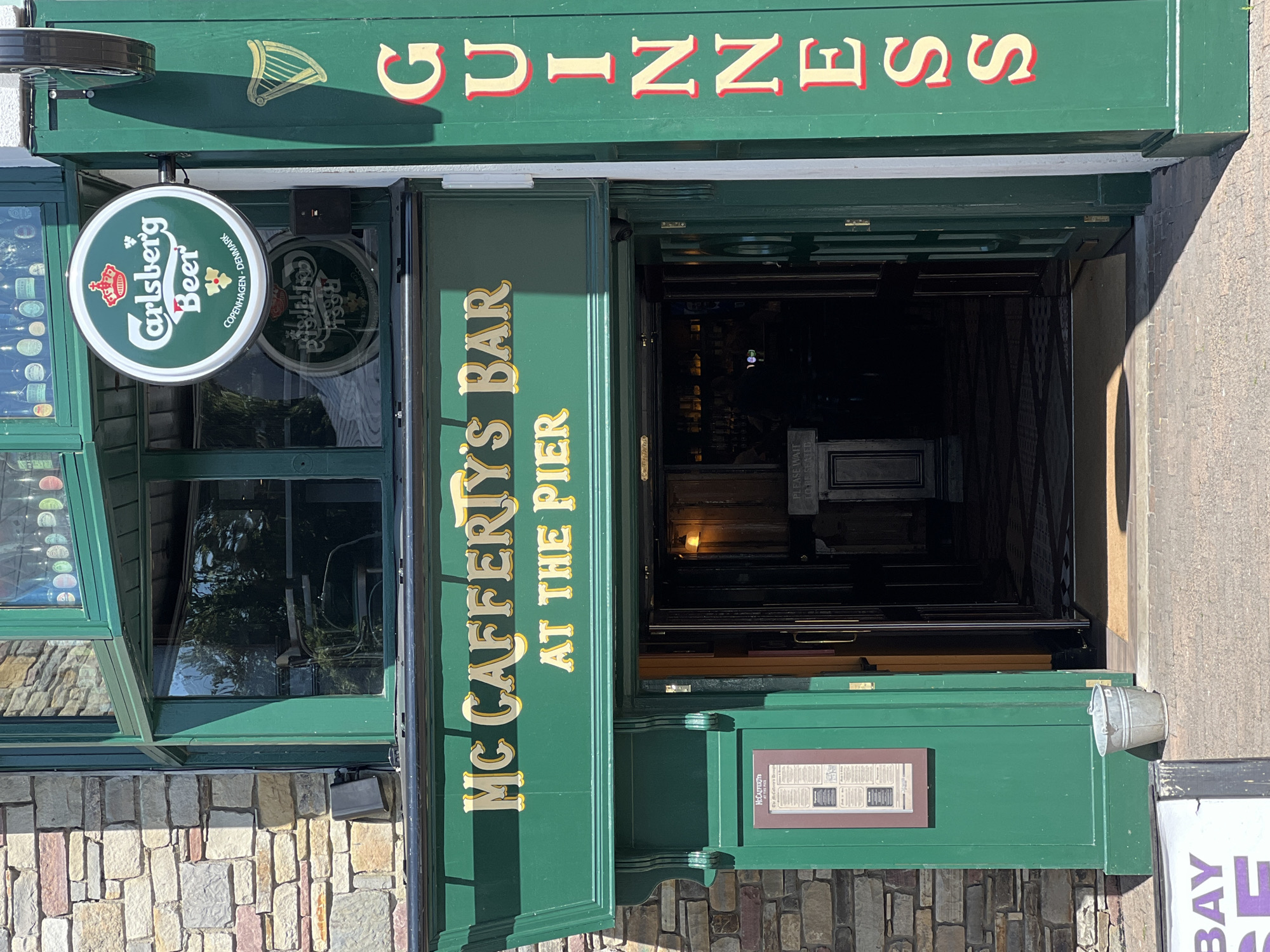 This is a full-length outdoor, eye-level, medium shot of McCafferty's Bar at The Pier in Donegal, Ireland. The bar is a two-story establishment with stone and green painted facades. A prominent green sign with gold lettering reads "MC CAFFERTY'S BAR AT THE PIER" above the entrance. To the right, a vertical green sign with gold lettering reads "GUINNESS." A circular Carlsberg Beer sign is affixed to the upper left facade.
The entrance to the bar is open, revealing a dimly lit interior with a bar and bottles visible. Several people, appearing as silhouettes, are inside the bar. To the left of the entrance is a green door with a framed menu or poster. A metal bucket sits on the pavement to the left of the door. A partial view of a white sign with purple lettering is visible in the lower left corner of the image. The sun is casting shadows, indicating a bright, sunny day.