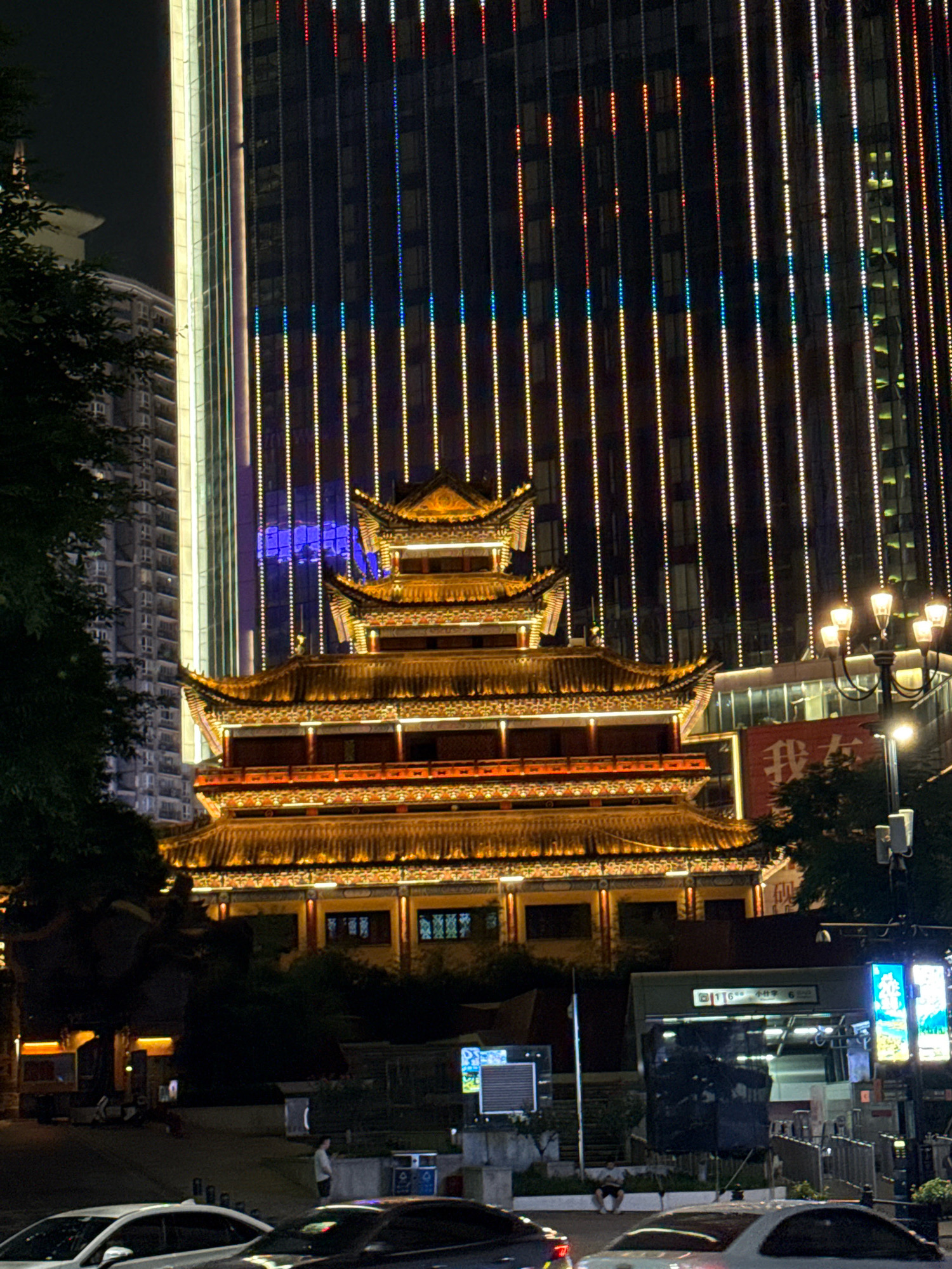 At night in Yuzhong District, China, a traditional multi-tiered Chinese pagoda-style building is illuminated with warm yellow lights. The pagoda has ornate, upturned eaves and red accents. Behind it, a modern skyscraper is adorned with vertical strands of colorful LED lights in red, blue, and yellow, creating a striking contrast. In the foreground, streetlights illuminate a road with cars, and several individuals are visible, some near a subway entrance. A red banner with Chinese characters is displayed on a building to the right of the pagoda. The scene captures a blend of historic architecture and contemporary urban development, characteristic of the Yuzhong District.
